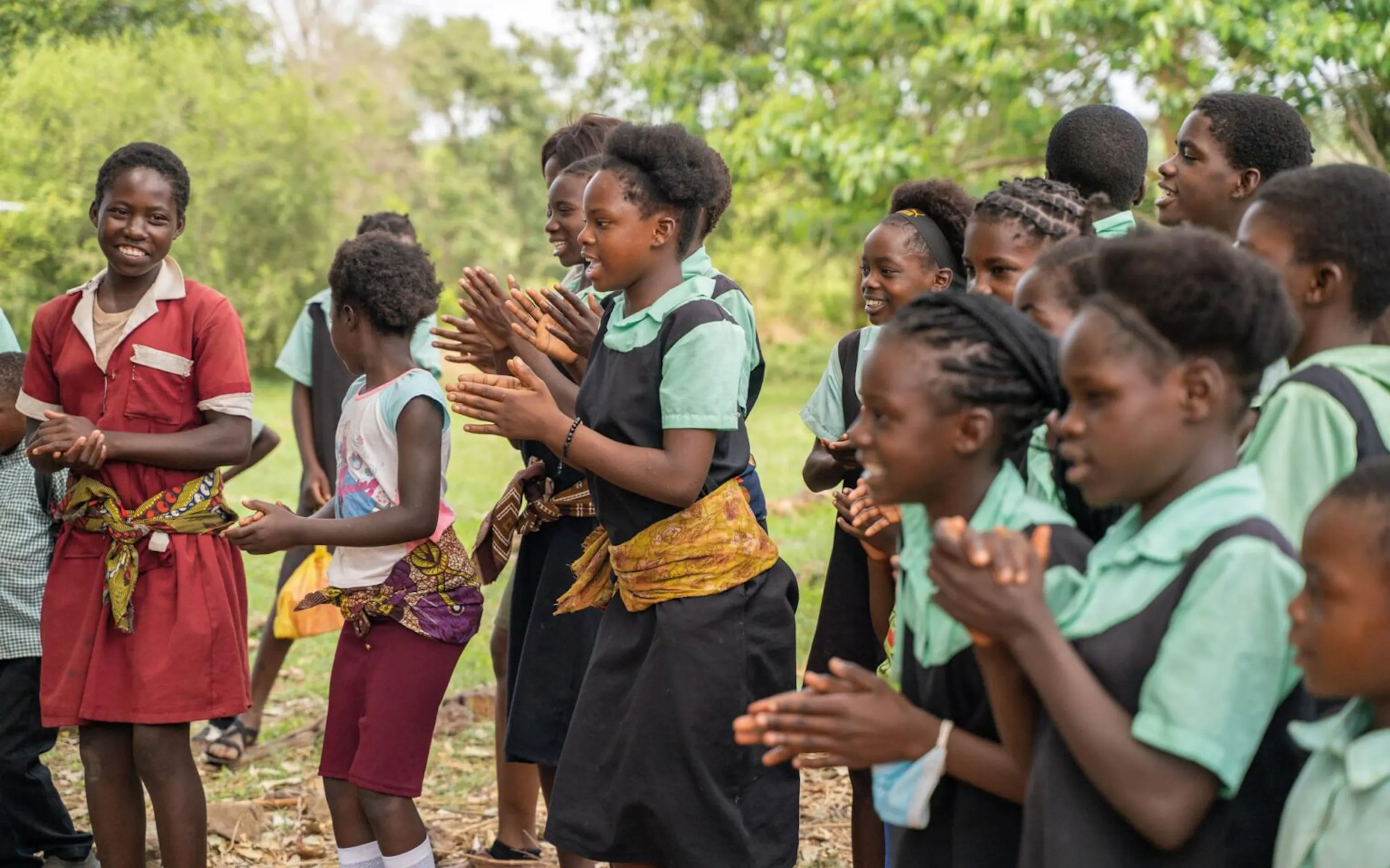 Schoolgirls in colorful uniforms clap and sing outdoors, gathered in a sunlit community scene with trees behind them.