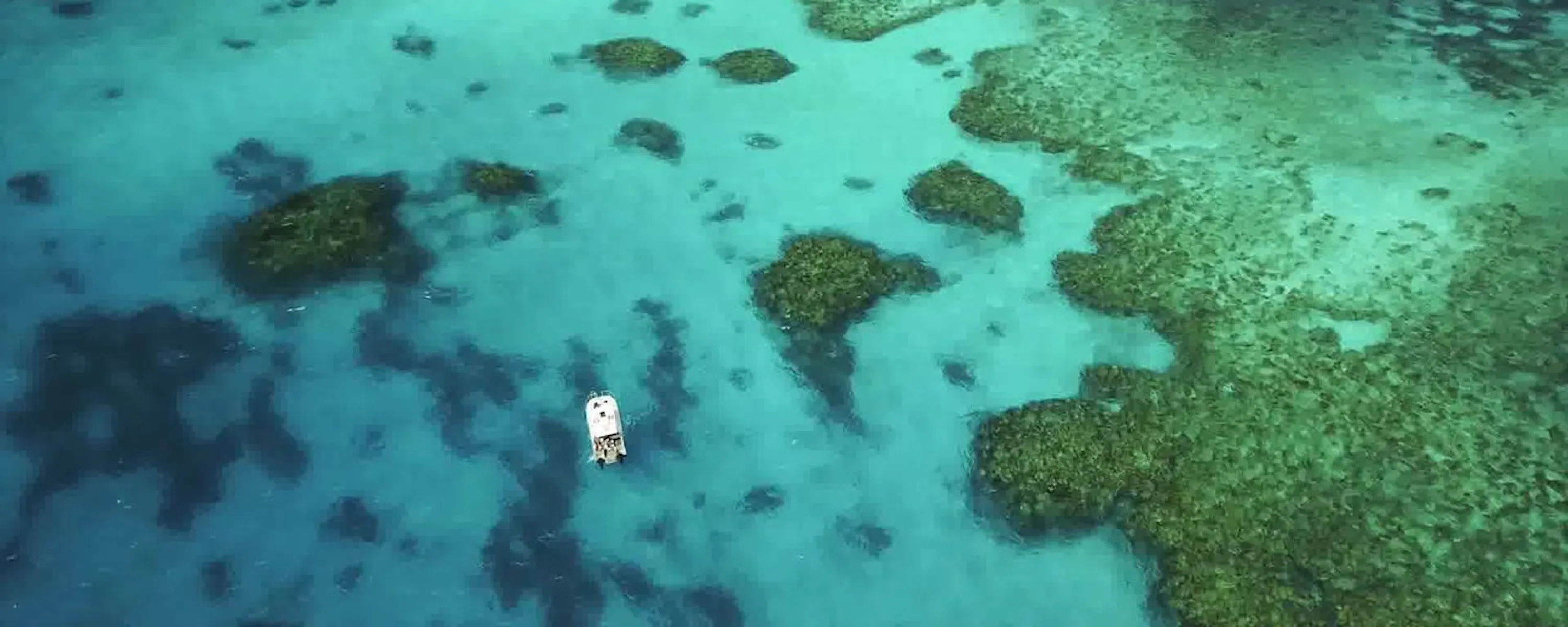 Aerial view of coral gardens and a narrow reef channel off the Great Barrier Reef, with a small boat below.