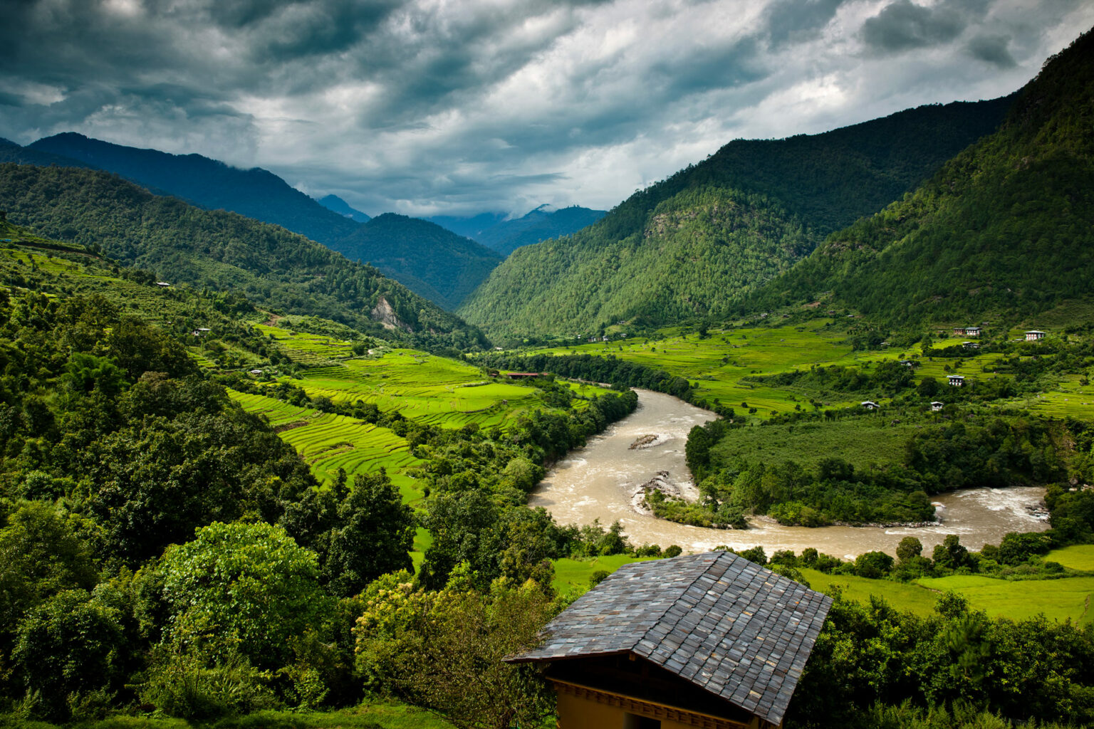 A river winds through Punakha valley below forested hills and terraced fields, with a retreat roof in the foreground.