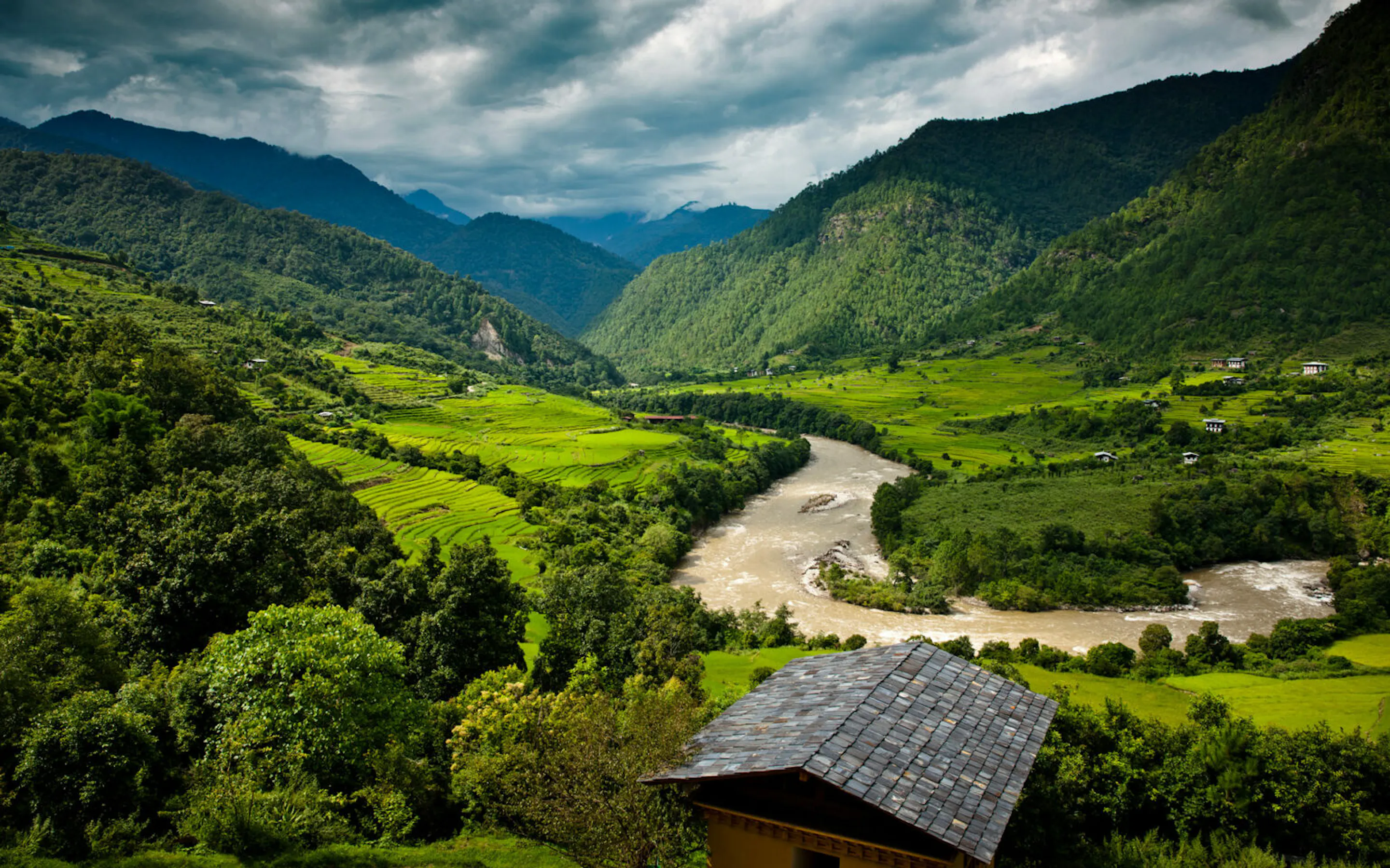 A river winds through Punakha valley below forested hills and terraced fields, with a retreat roof in the foreground.