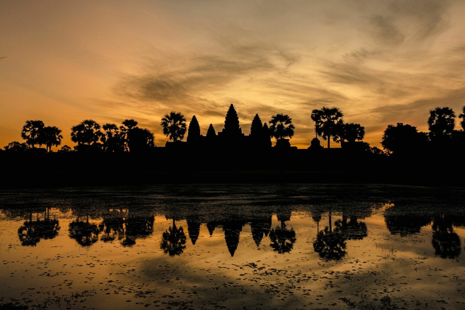 Temple towers silhouette against a glowing sky at Angkor Wat, their reflection mirrored in still water at sunset.