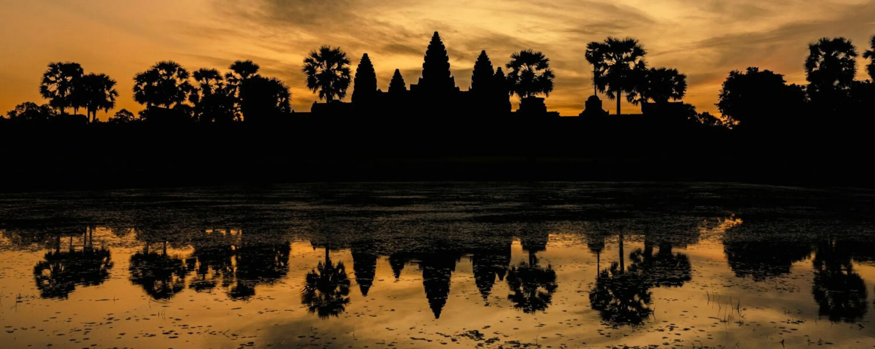 Temple towers silhouette against a glowing sky at Angkor Wat, their reflection mirrored in still water at sunset.