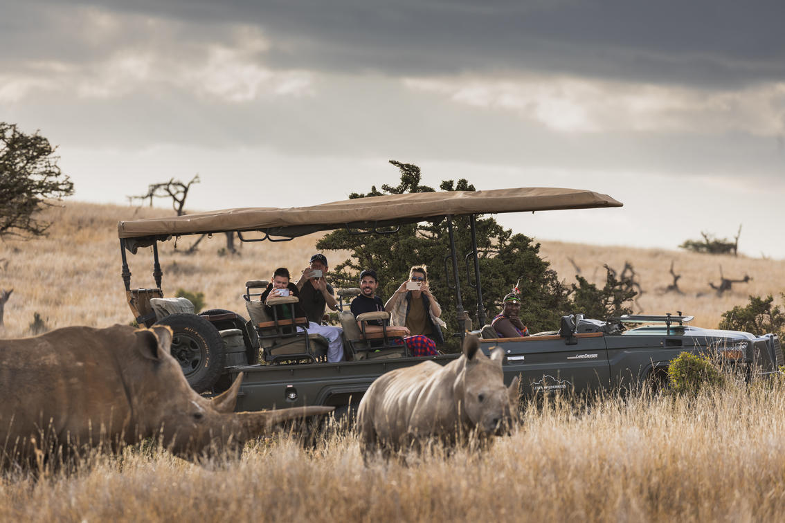 Safari guests photograph two rhinos from an open vehicle in dry grassland at Lewa, Kenya, under cloudy skies.