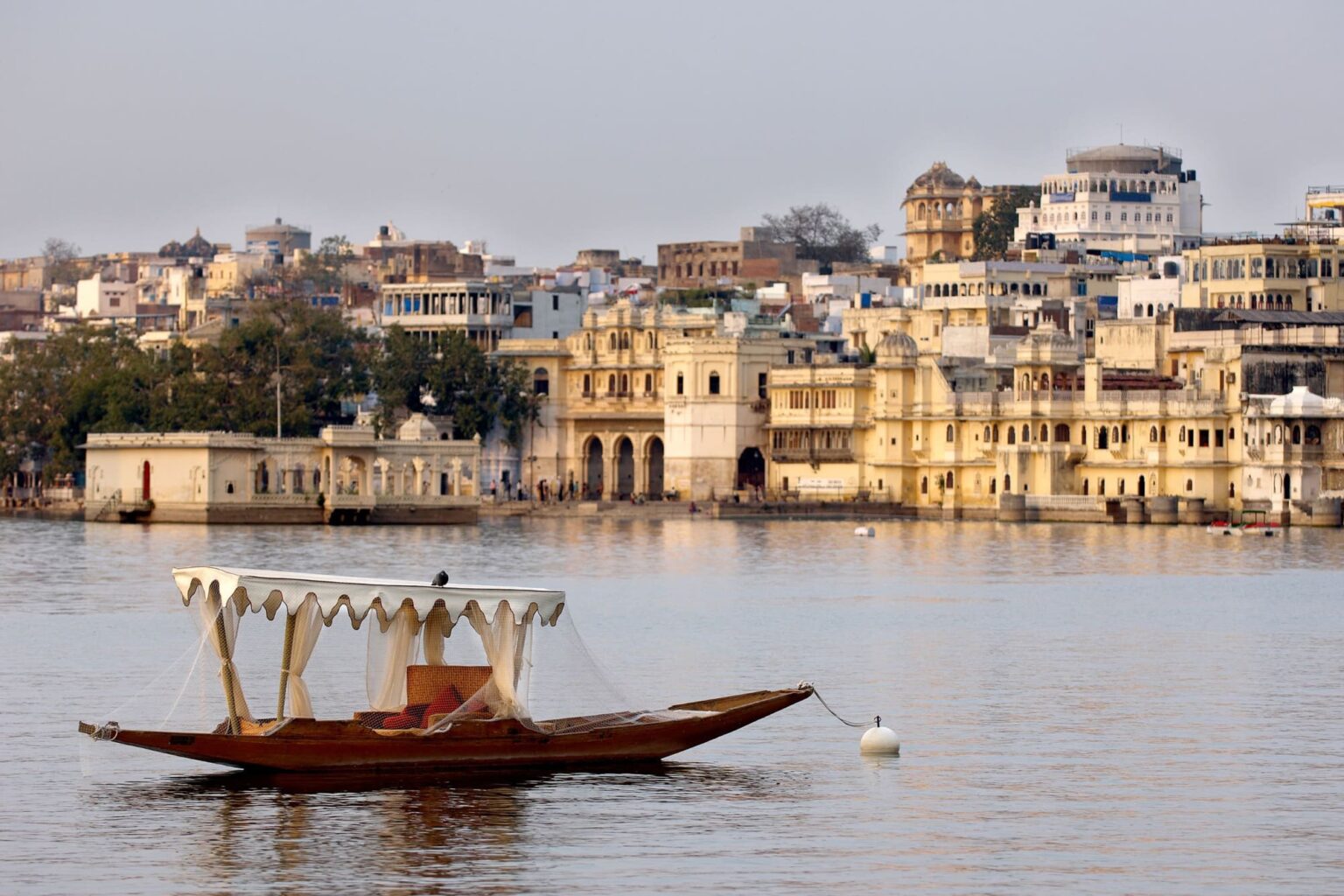 A wooden boat drifts on Lake Pichola, with palace-lined shores, layered hills, and the skyline of Udaipur behind it.