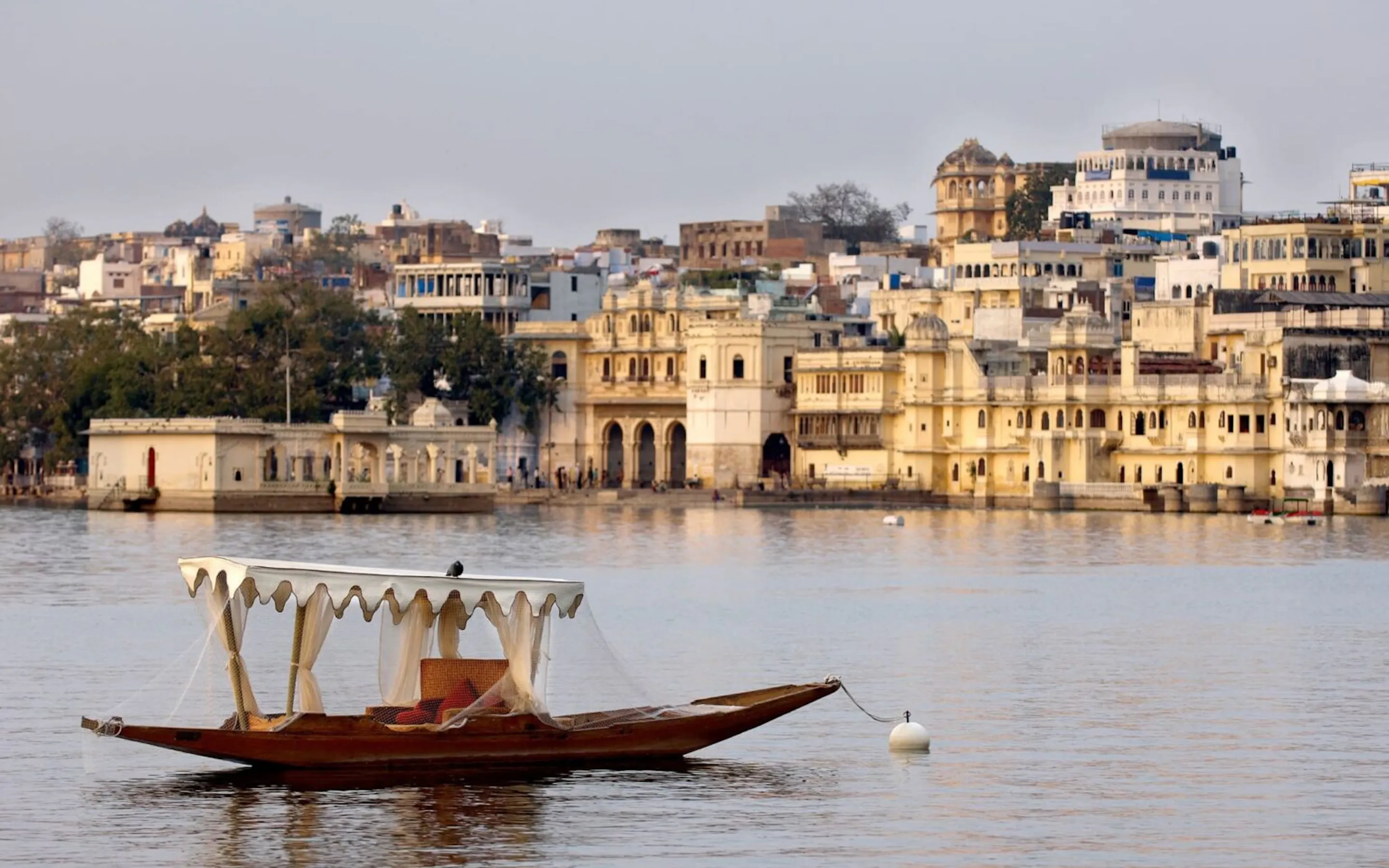 A wooden boat drifts on Lake Pichola, with palace-lined shores, layered hills, and the skyline of Udaipur behind it.