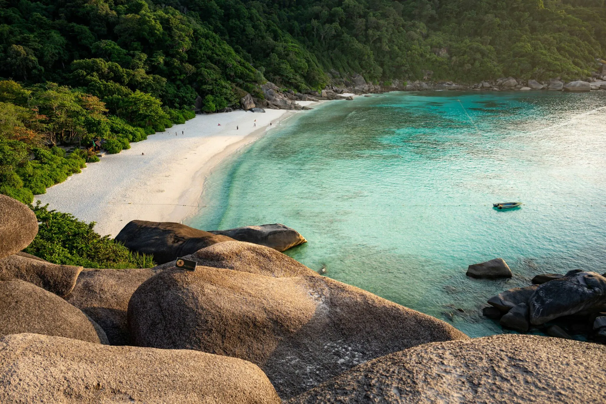 Turquoise water laps a crescent beach below smooth granite boulders, with forested hills framing the bay.