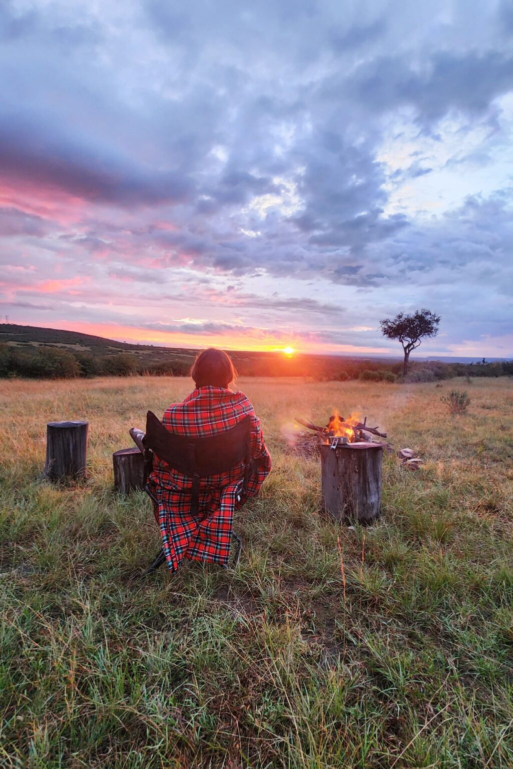 A traveler wrapped in a shuka watching sunset beside a campfire against the backdrop of Kenya's Maasai Mara.
