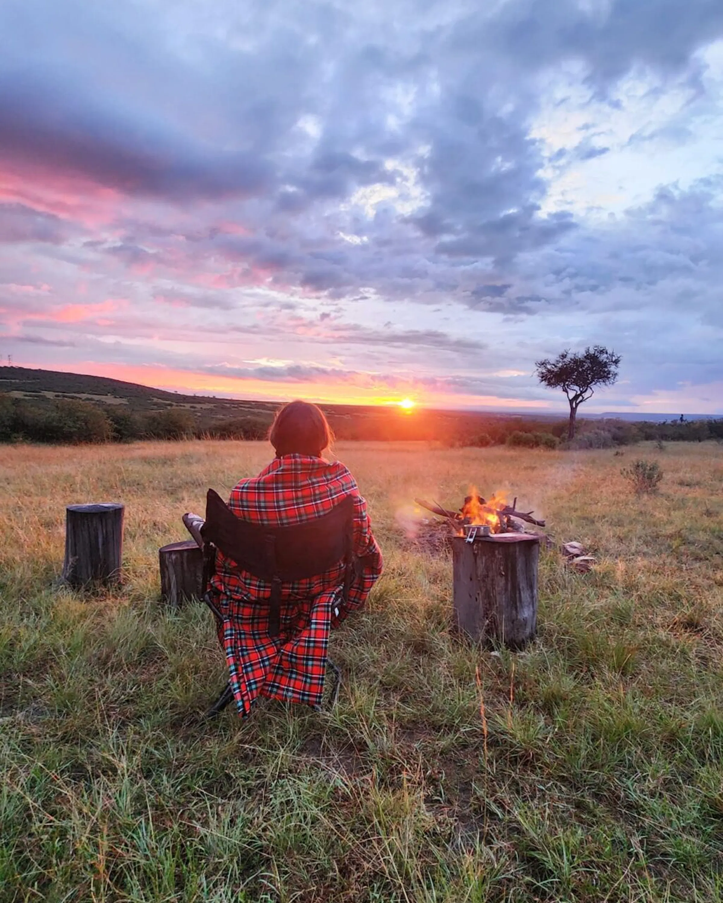 A traveler wrapped in a shuka watching sunset beside a campfire against the backdrop of Kenya's Maasai Mara.