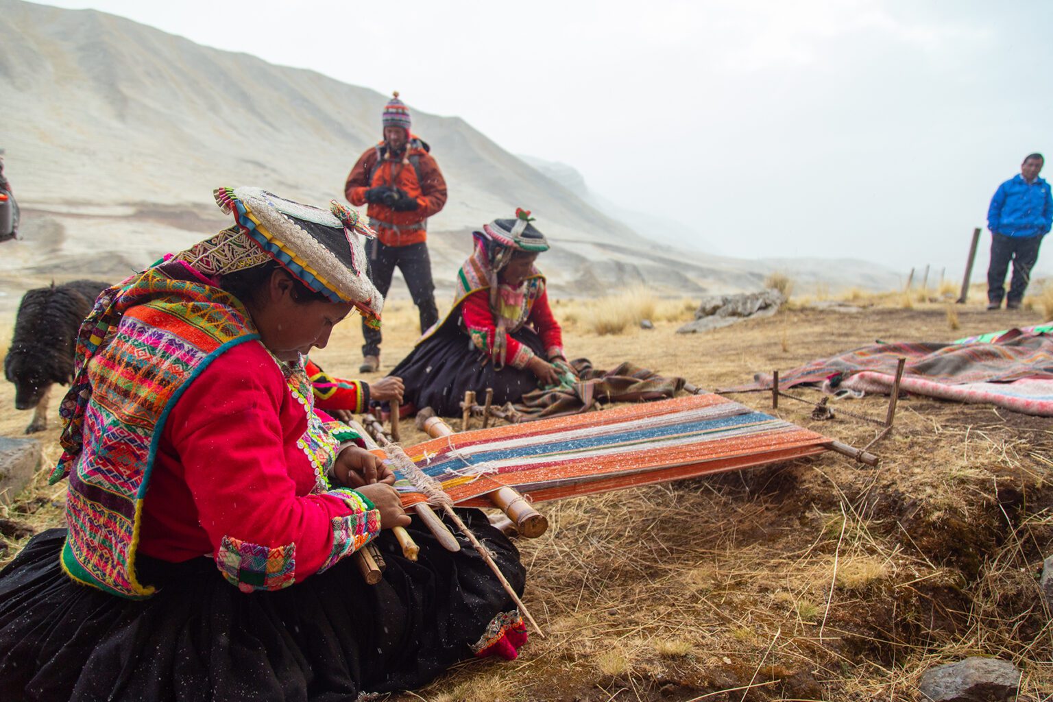Two women weaving colorful textiles in a windswept Andean village against the backdrop of the Peruvian Andes.