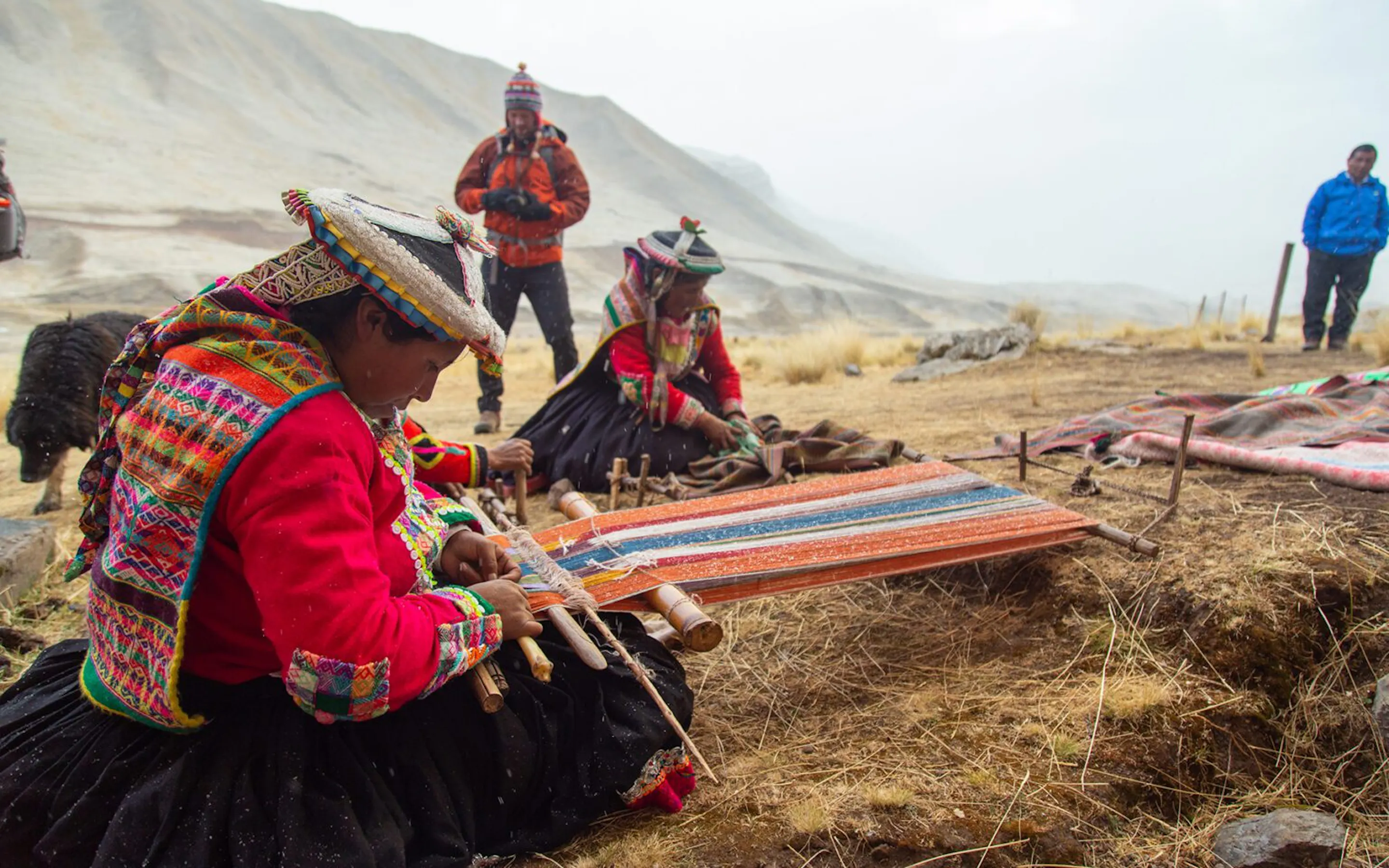 Two women weaving colorful textiles in a windswept Andean village against the backdrop of the Peruvian Andes.