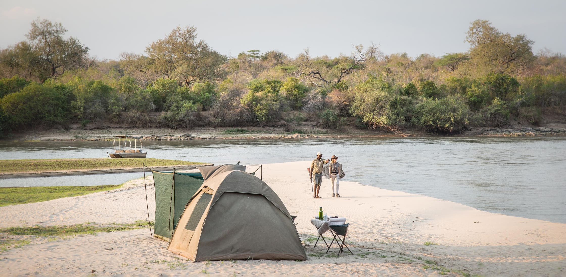 Fly-camping tents line a pale sandbank in Nyerere, with the river, distant walkers, and woodland stretching beyond.