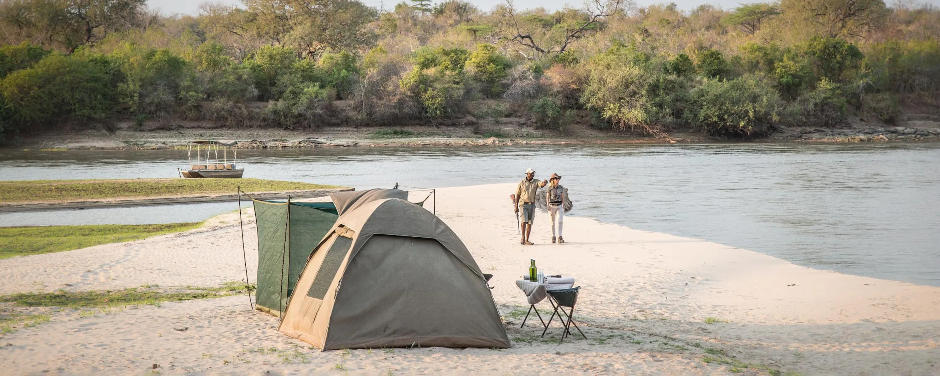 Fly-camping tents line a pale sandbank in Nyerere, with the river, distant walkers, and woodland stretching beyond.