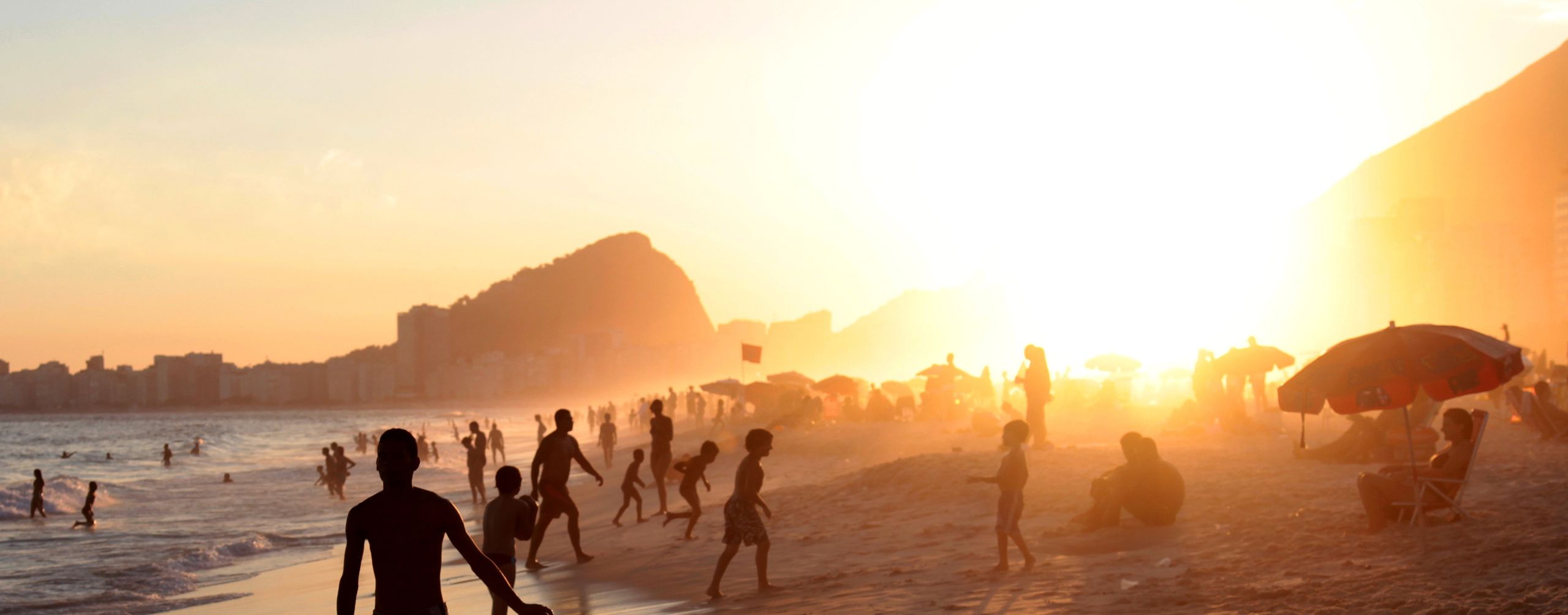 Silhouetted beachgoers walk along the shoreline in Rio, with warm sunset light, surf, and city cliffs beyond.