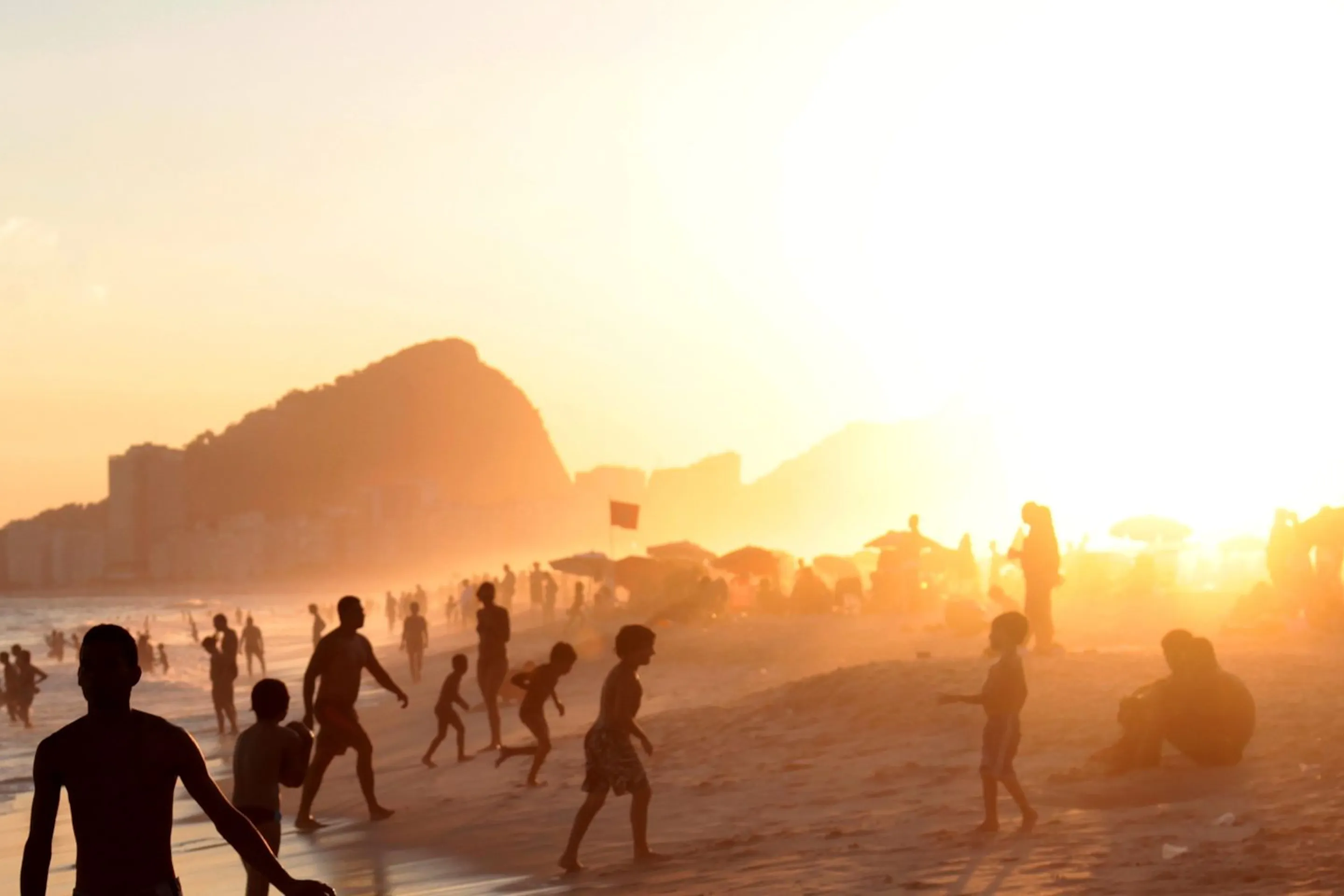 Silhouetted beachgoers walk along the shoreline in Rio, with warm sunset light, surf, and city cliffs beyond.