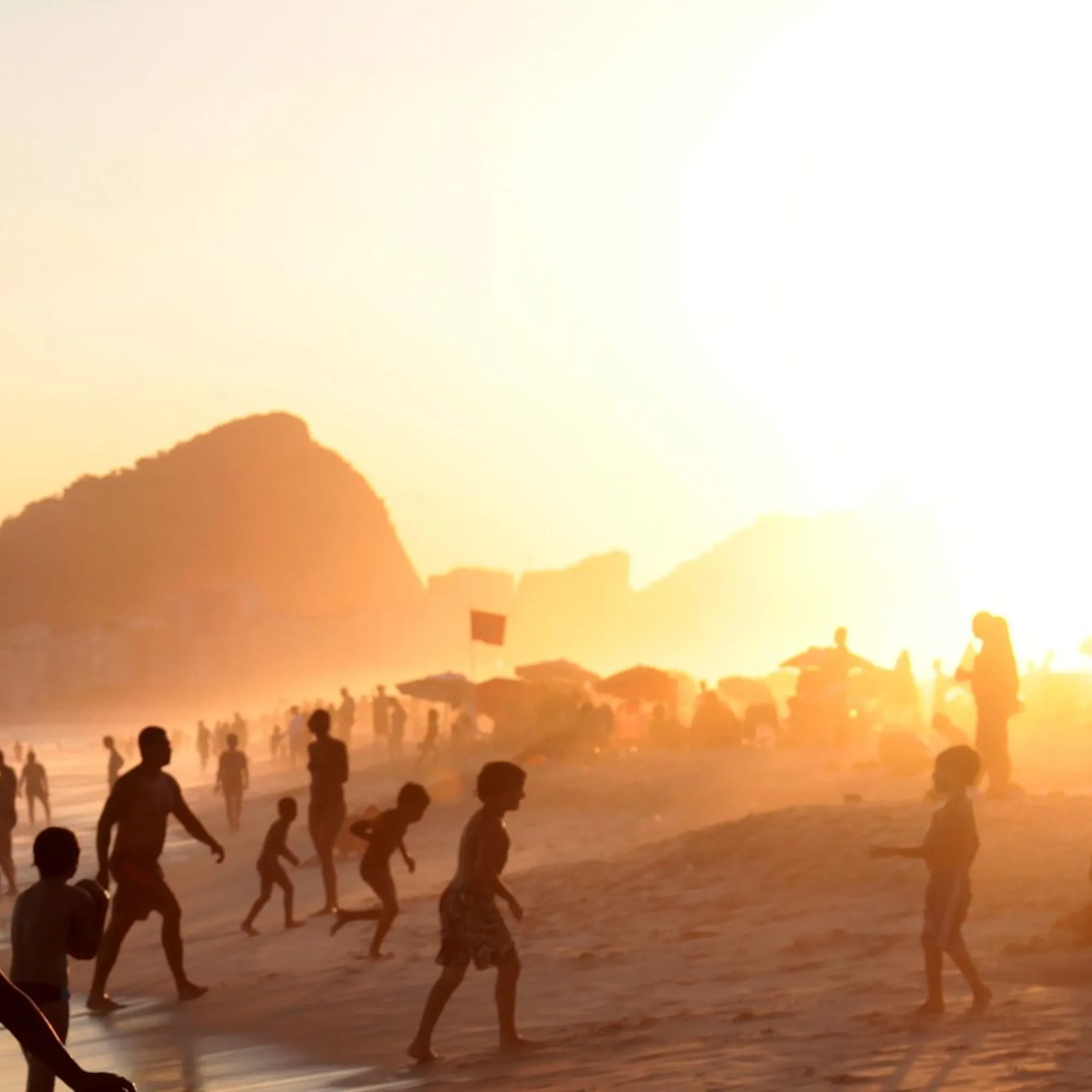 Silhouetted beachgoers walk along the shoreline in Rio, with warm sunset light, surf, and city cliffs beyond.