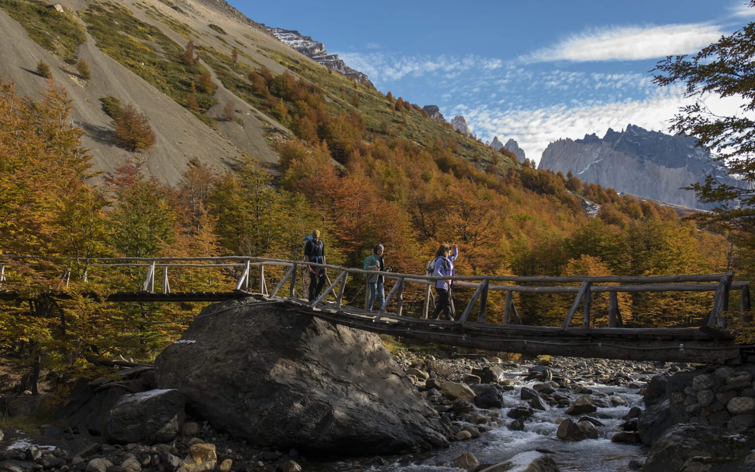 Hikers cross a bridge above a river inTorres del Paine with jagged peaks and open slopes beyond.