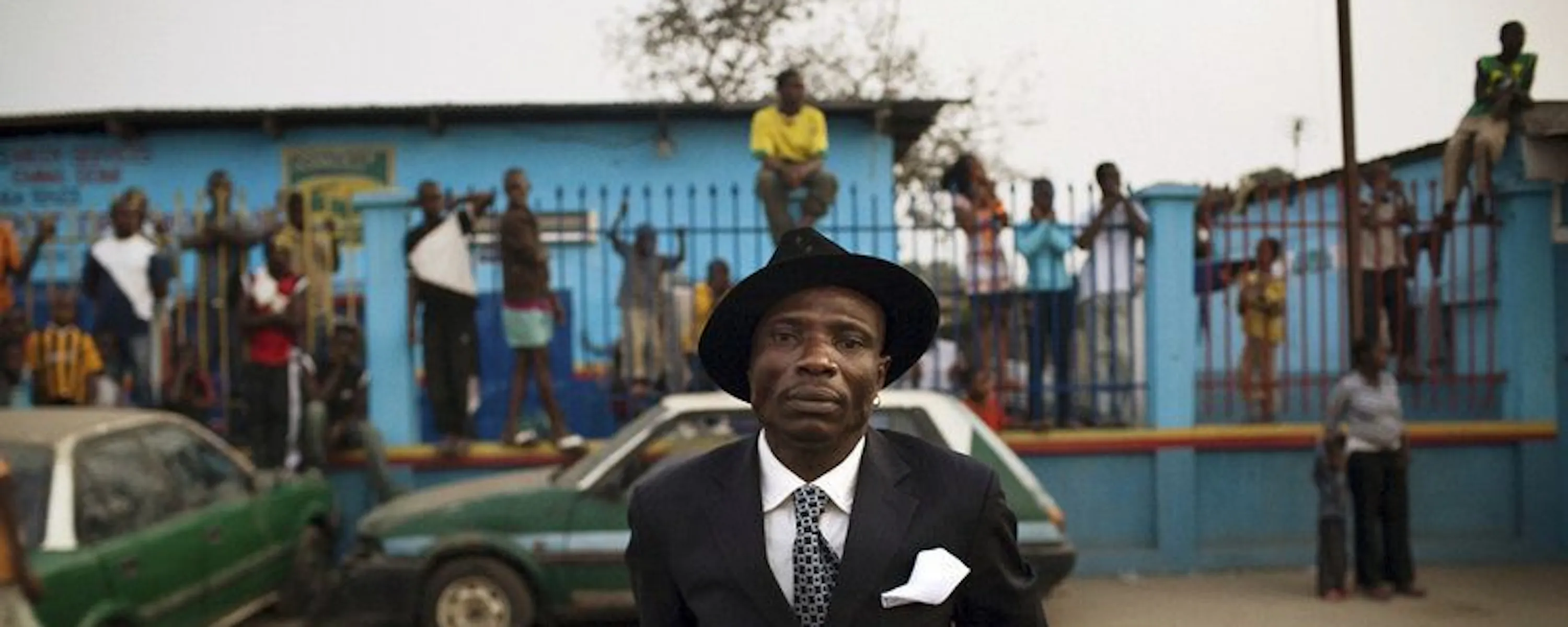 A sharply dressed sapeur stands before a lively Brazzaville street scene, framed by friends and bright color.