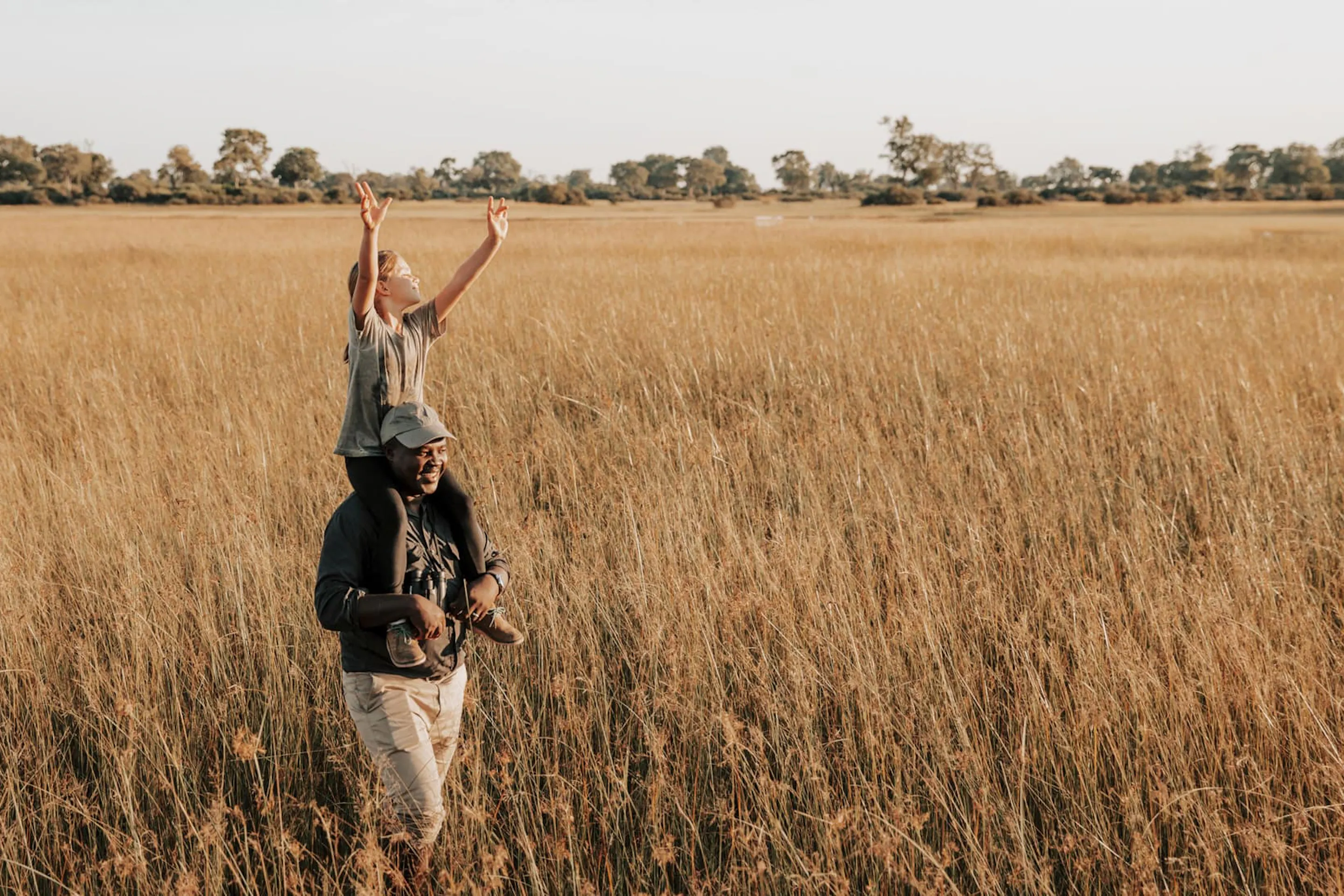 A child stands in tall Okavango grass, holding a branch high while a guide watches nearby under open sky.