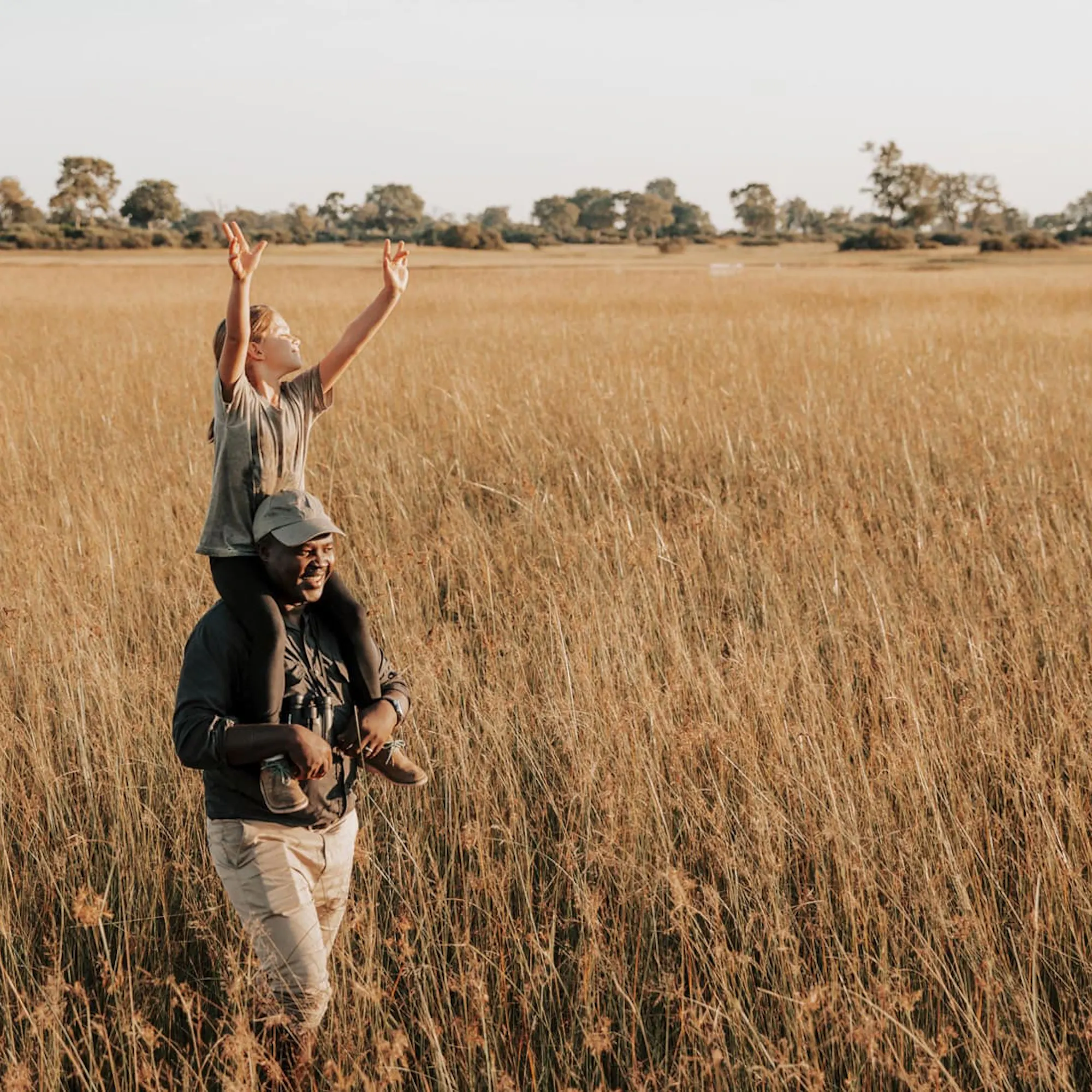 A child stands in tall Okavango grass, holding a branch high while a guide watches nearby under open sky.