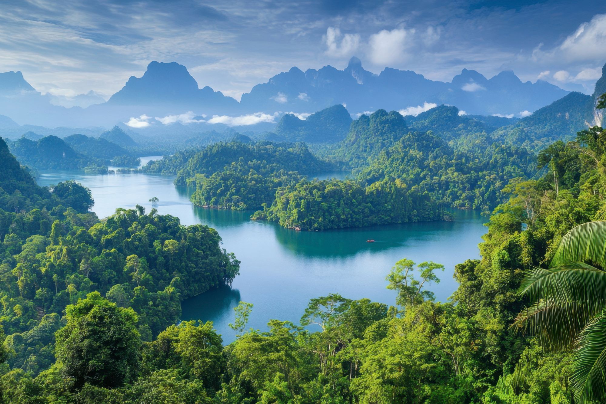 An emerald lake winds through dense jungle, with limestone peaks rising beyond layered, misty ridgelines in the distance.