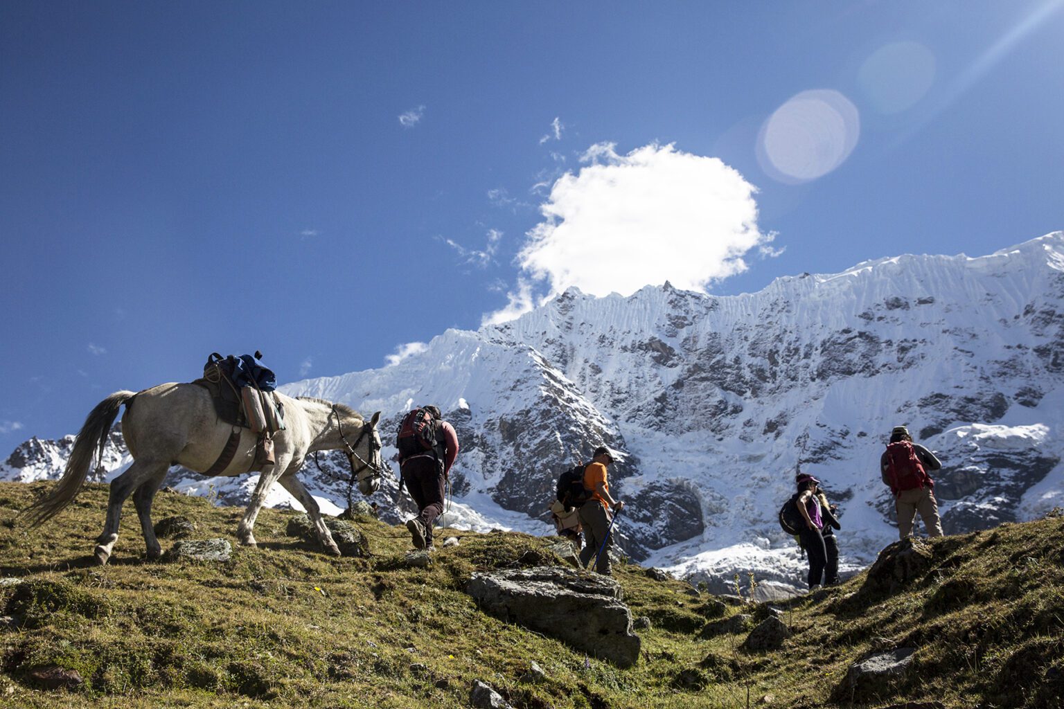 Hikers and a packhorse cross rocky grassland below snow-covered peaks on the Salkantay Trek near Cusco, Peru.