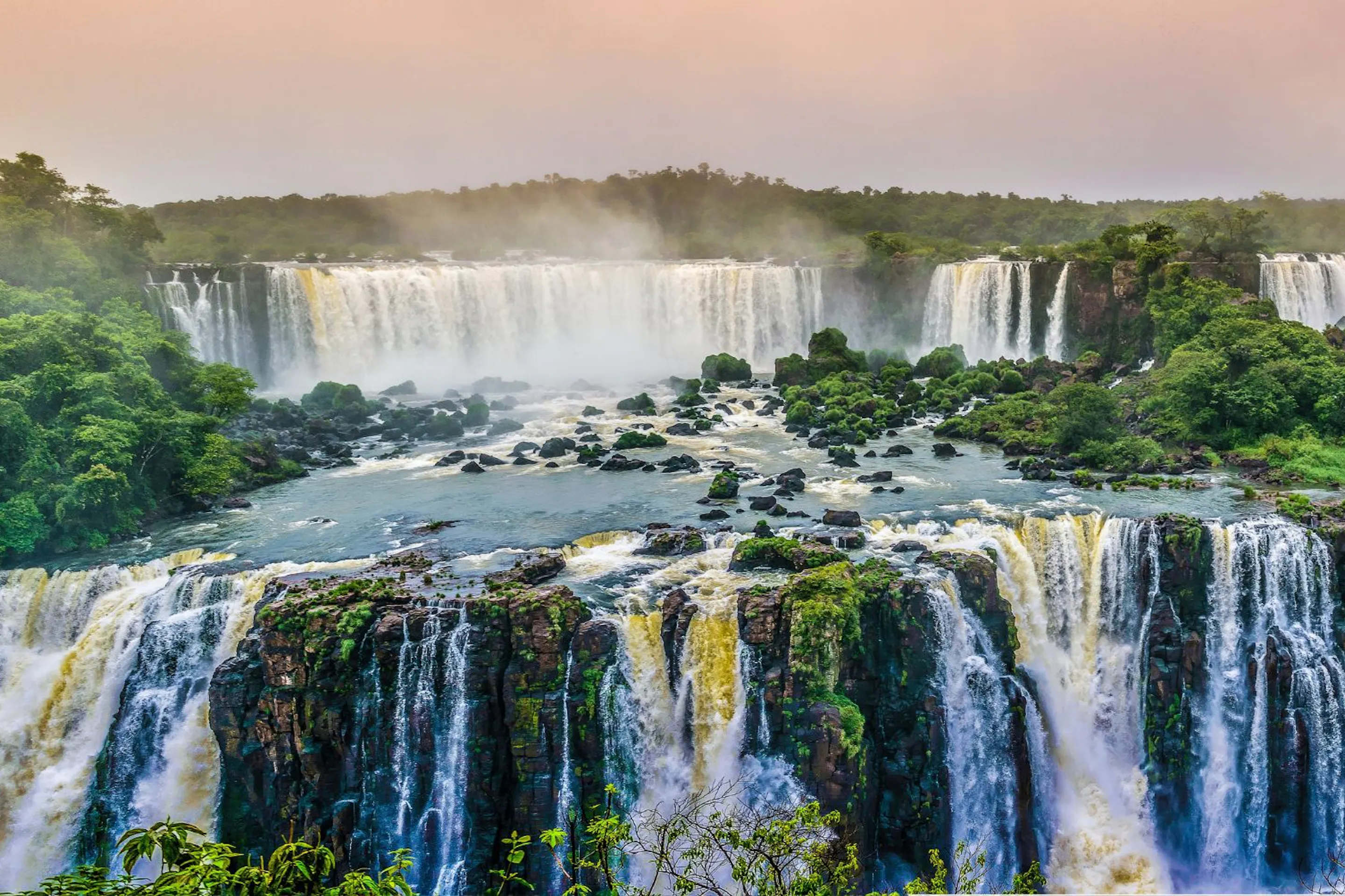 Powerful white cascades thunder through dense green forest at Iguacu Falls, viewed from above the deep gorge below.