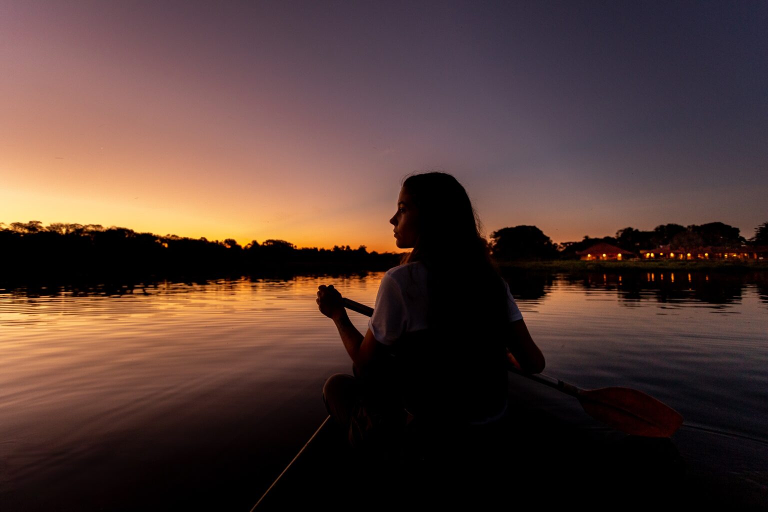 A girl paddles a canoe across still water at dusk near Caiman, with Brazil's Pantanal fading into shadow.