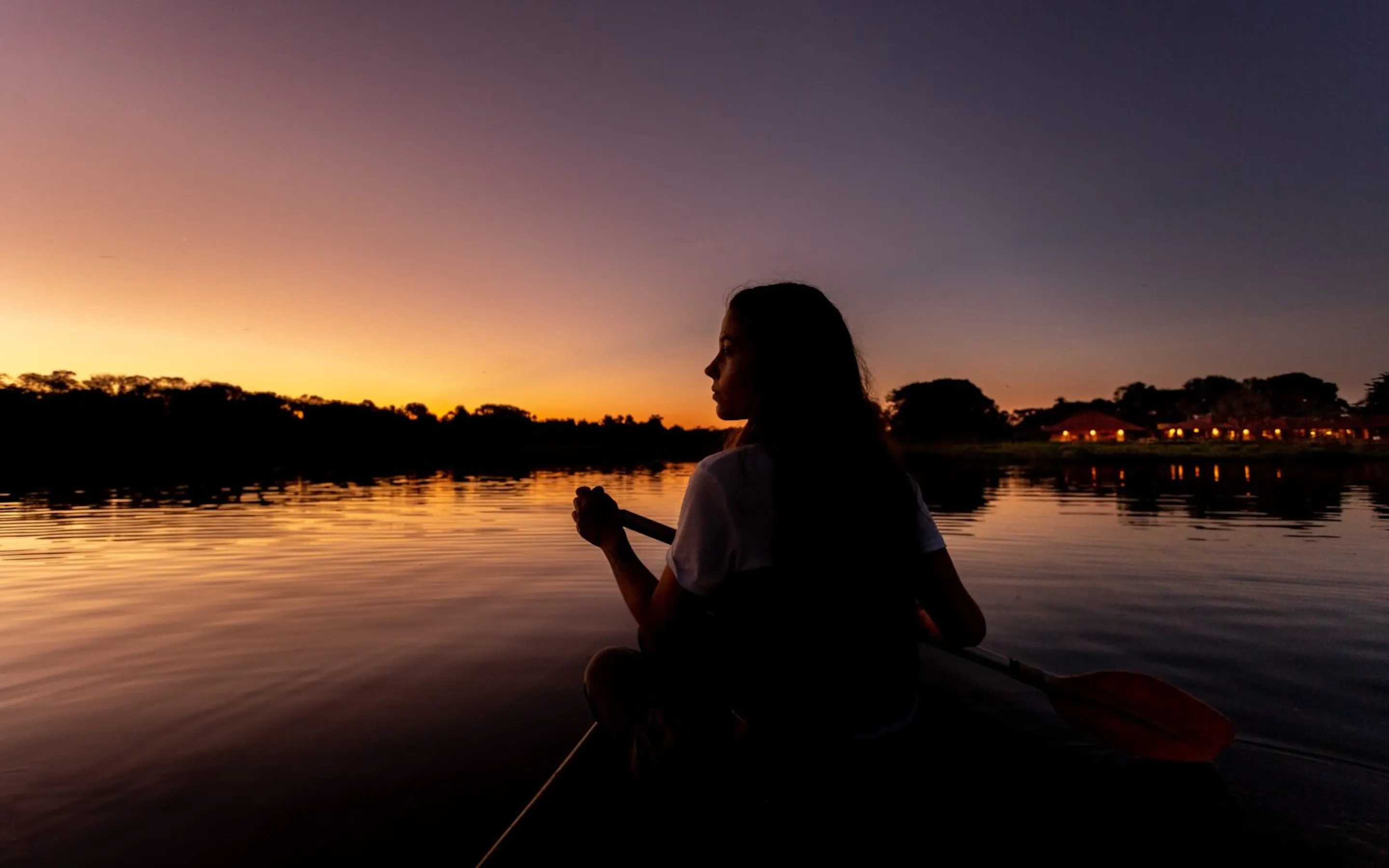 A girl paddles a canoe across still water at dusk near Caiman, with Brazil's Pantanal fading into shadow.