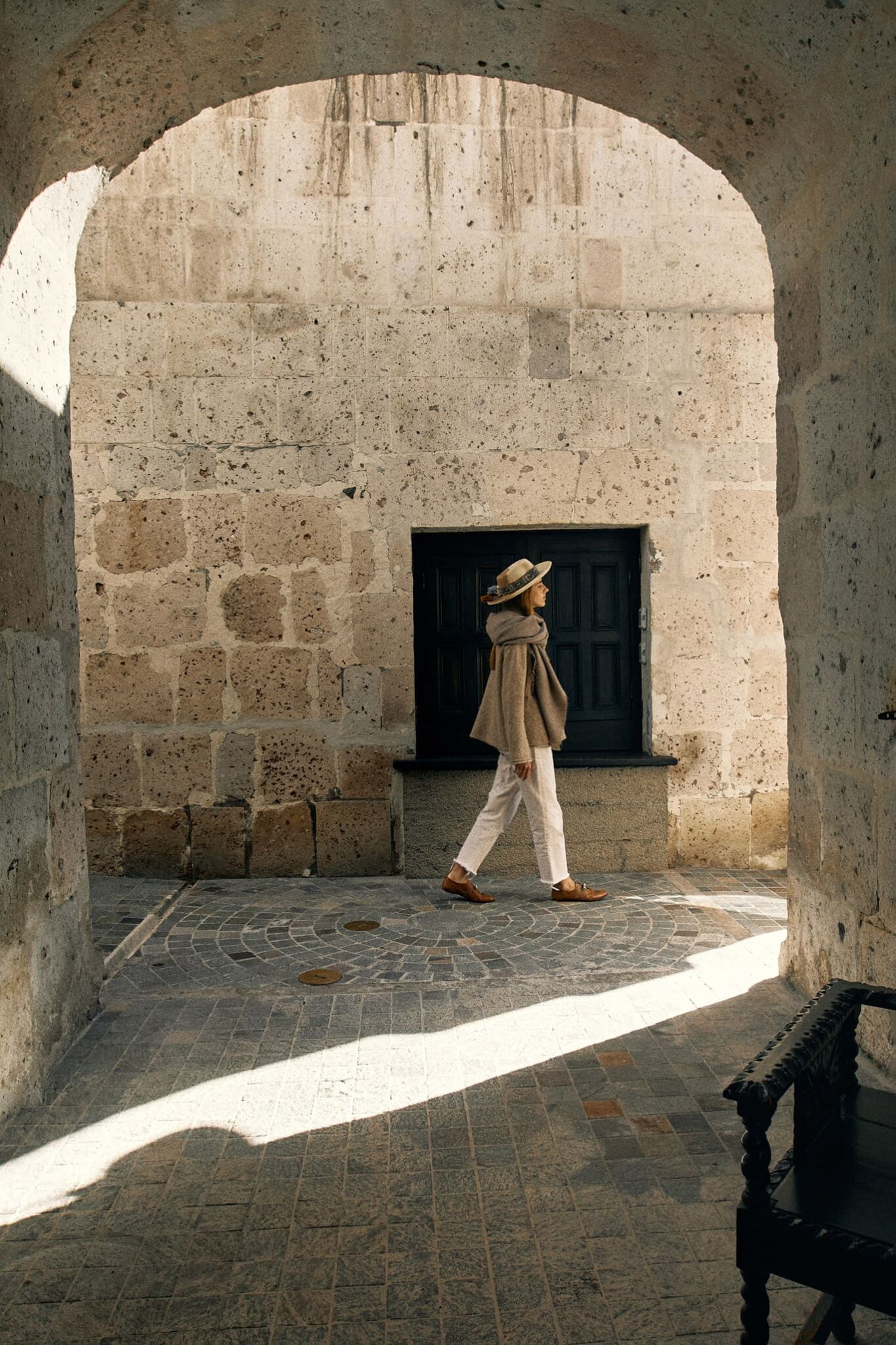 A woman walks beneath stone arches in Arequipa, with strong sunlight cutting across the white volcanic walls.