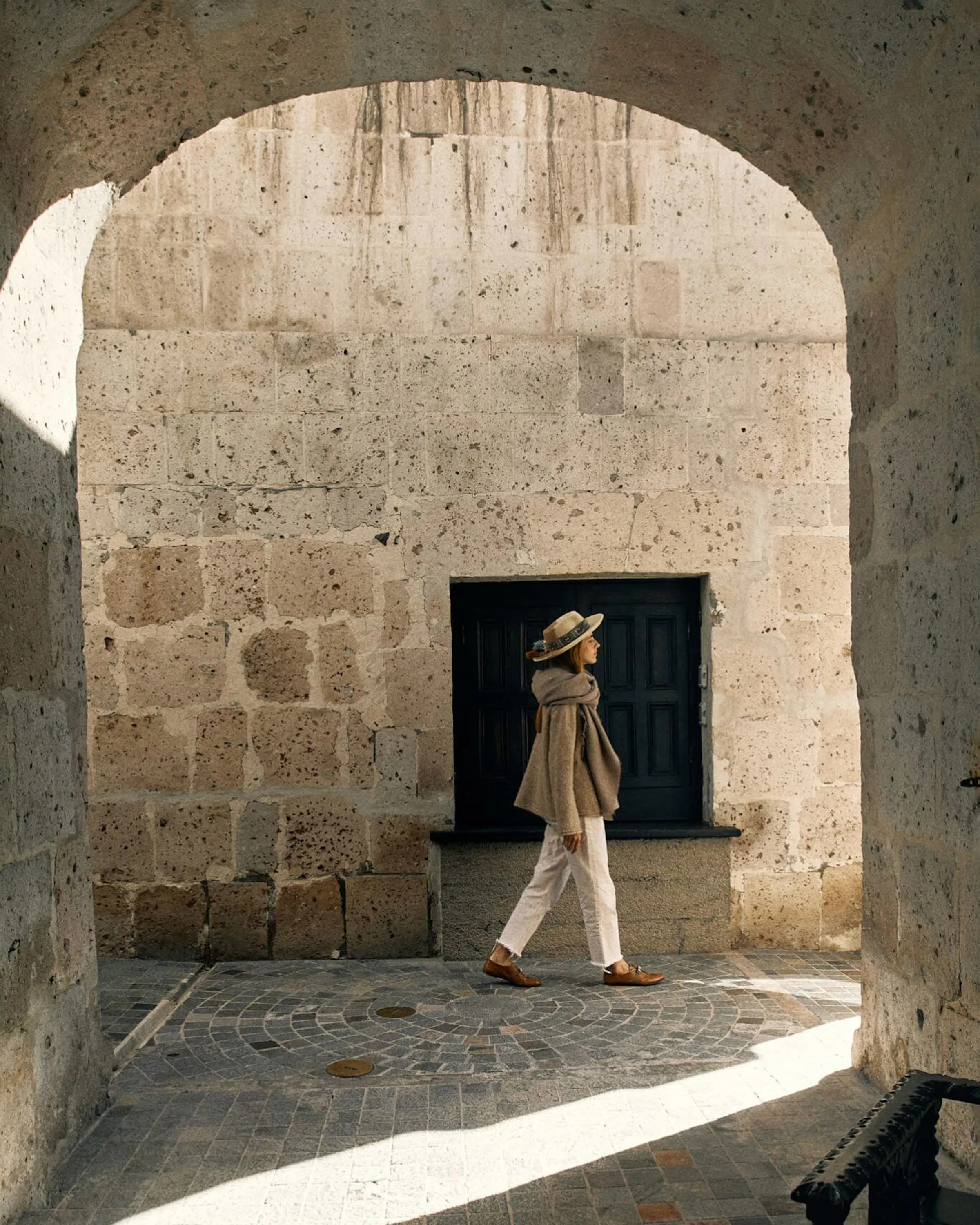 A woman walks beneath stone arches in Arequipa, with strong sunlight cutting across the white volcanic walls.