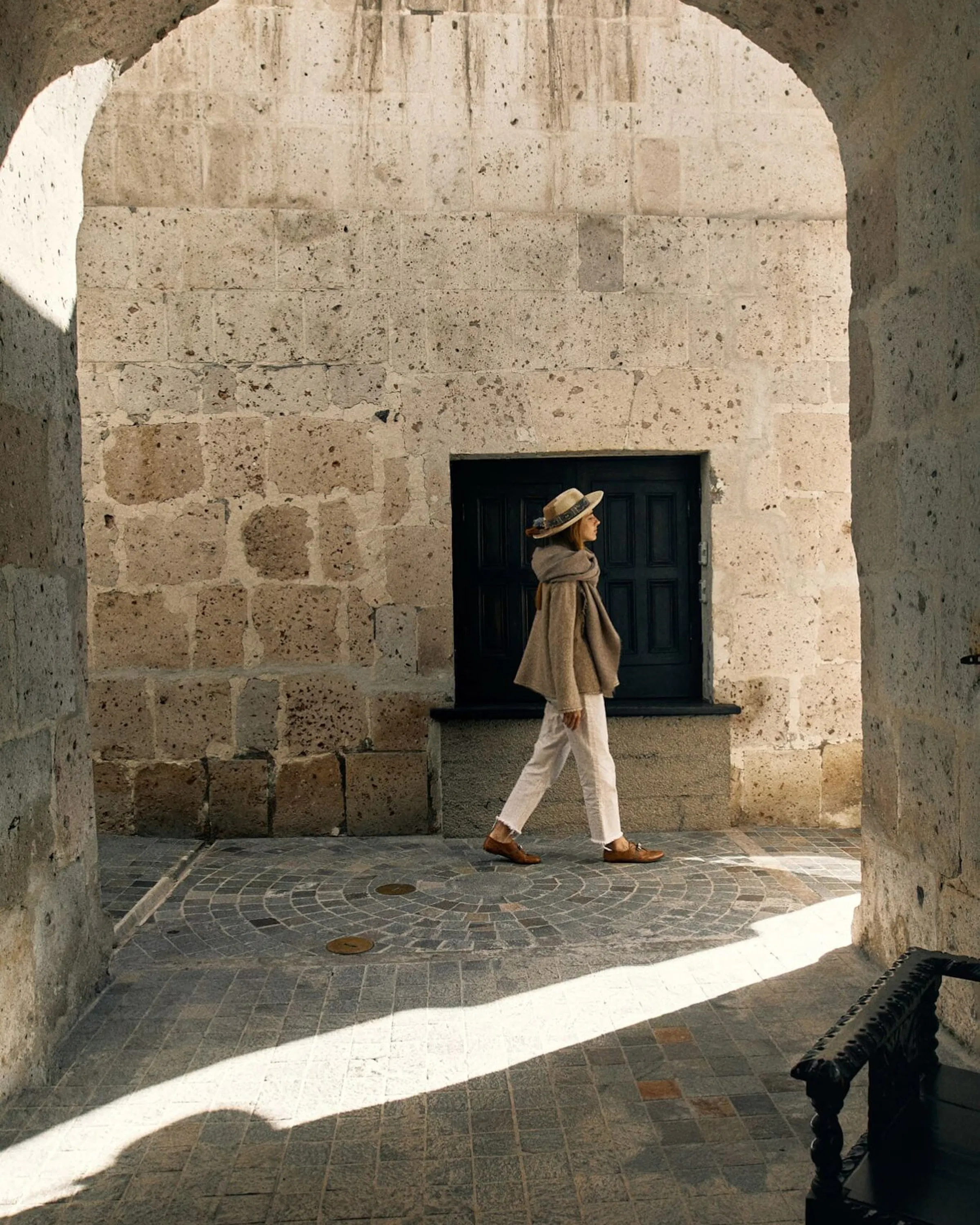 A woman walks beneath stone arches in Arequipa, with strong sunlight cutting across the white volcanic walls.
