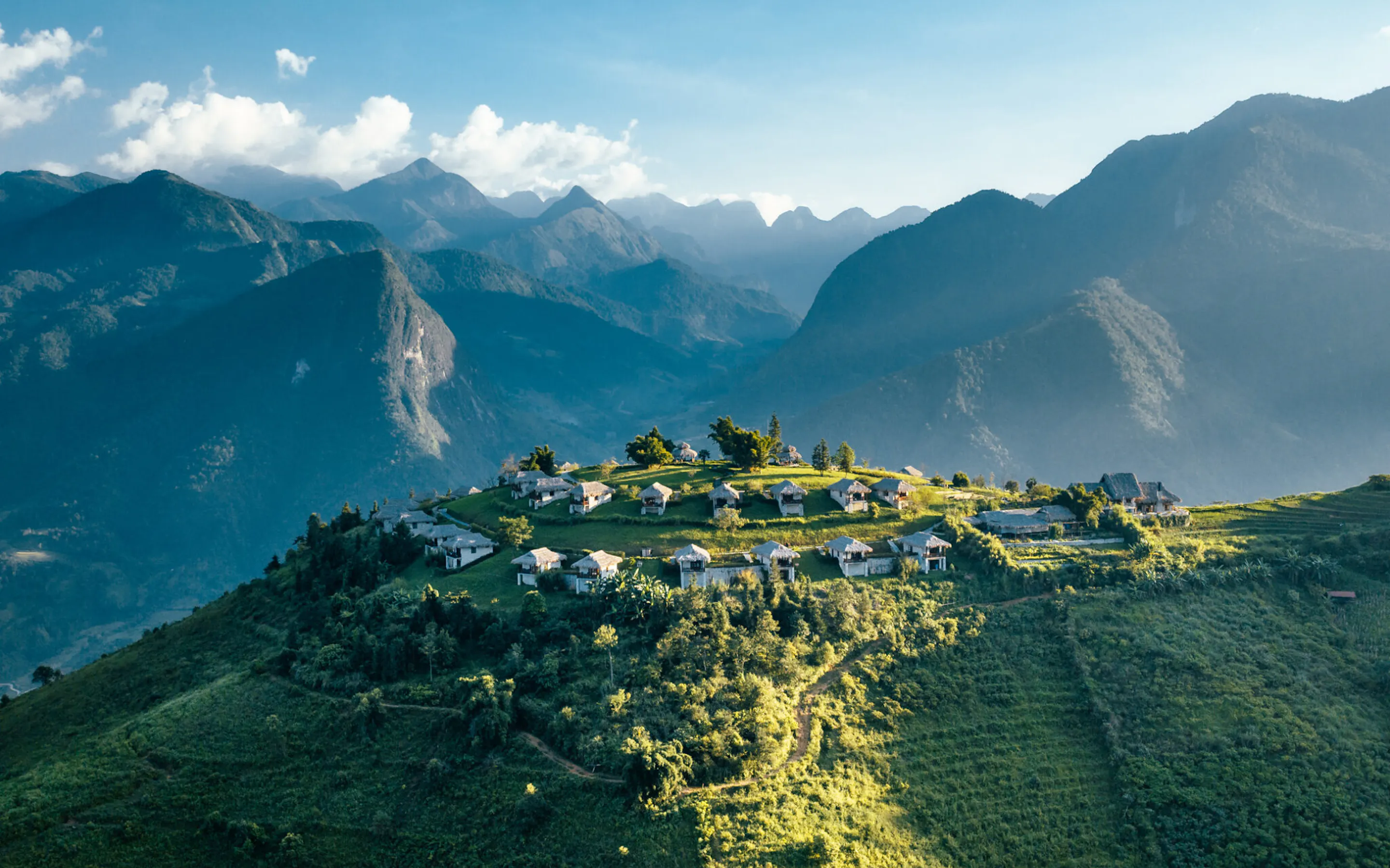 Mountain ridges frame a hilltop lodge complex with scattered villas and paths, lit by afternoon sun and drifting clouds.