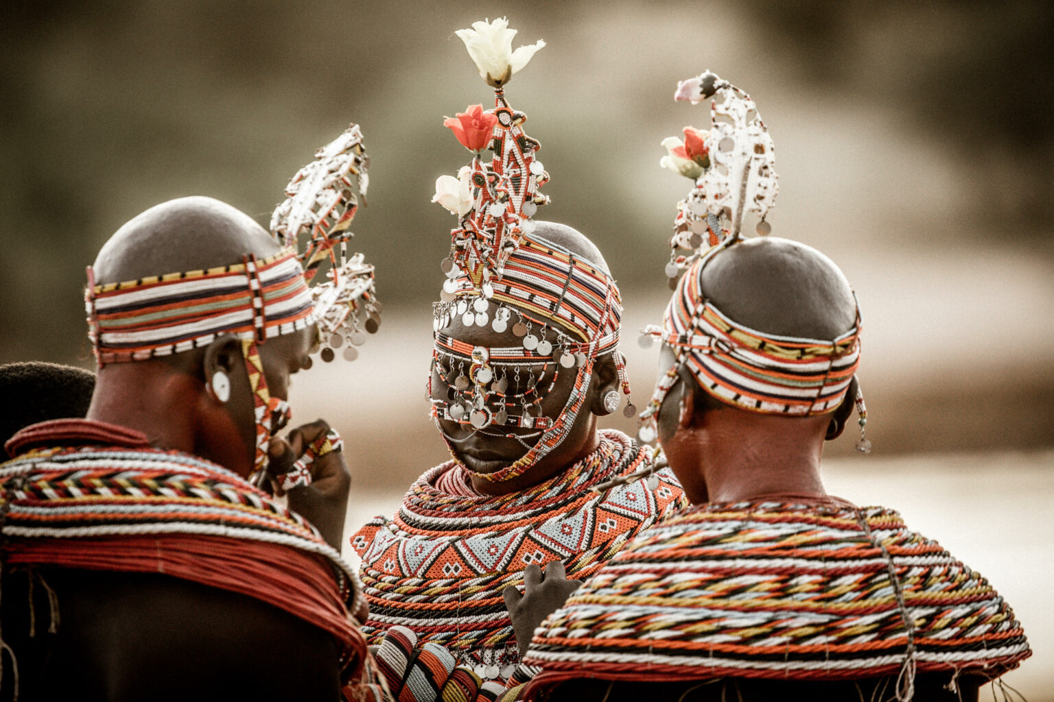 Three Samburu women are seen from behind in beaded dress in Kenya's Samburu, walking across open ground together.