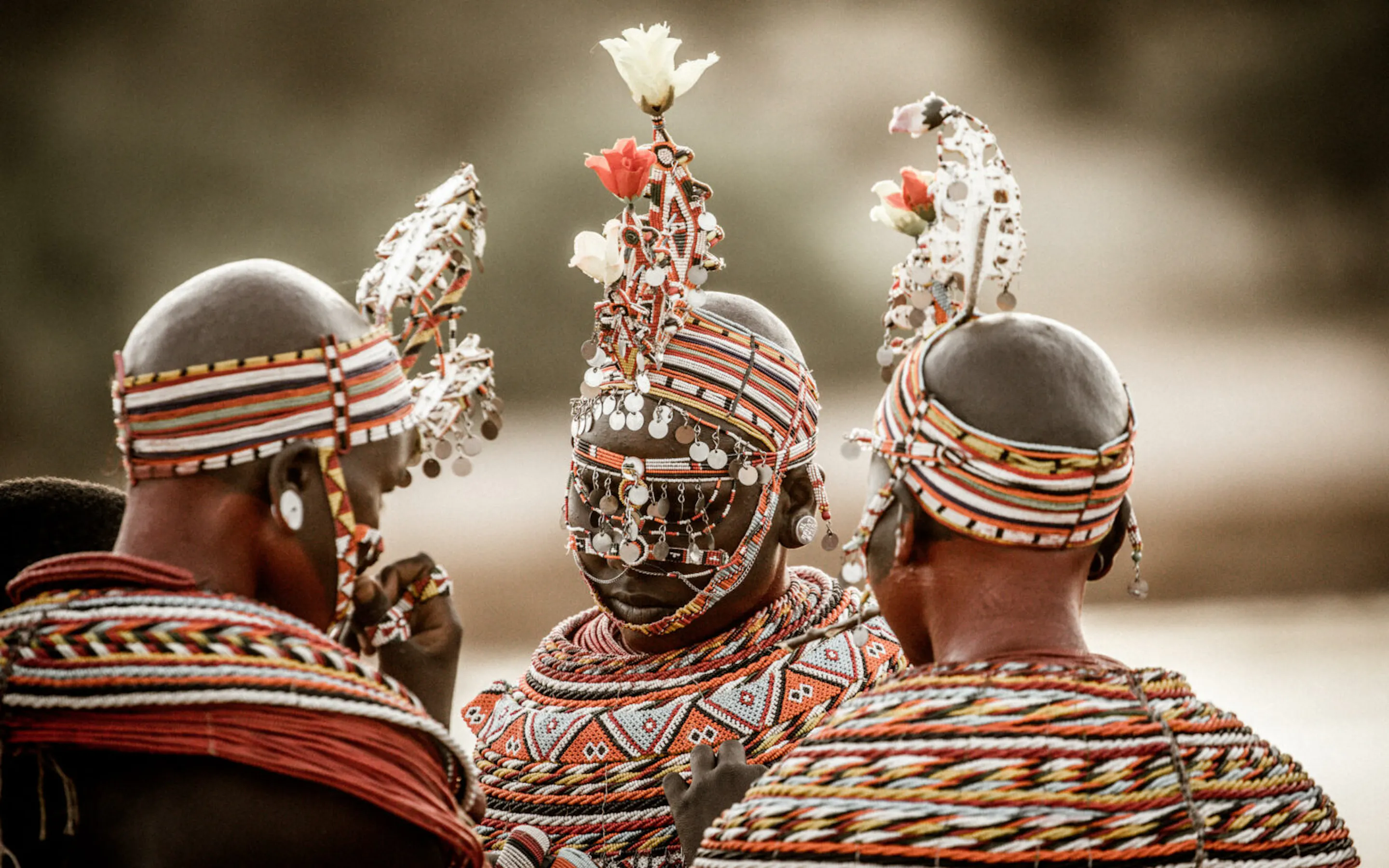 Three Samburu women are seen from behind in beaded dress in Kenya's Samburu, walking across open ground together.