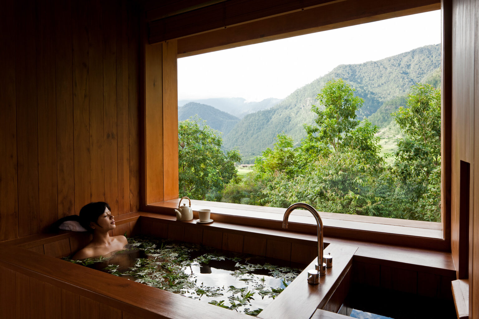 A woman relaxes in a deep tub at COMO Uma Punakha, framed by wood paneling and misty hills beyond the window.