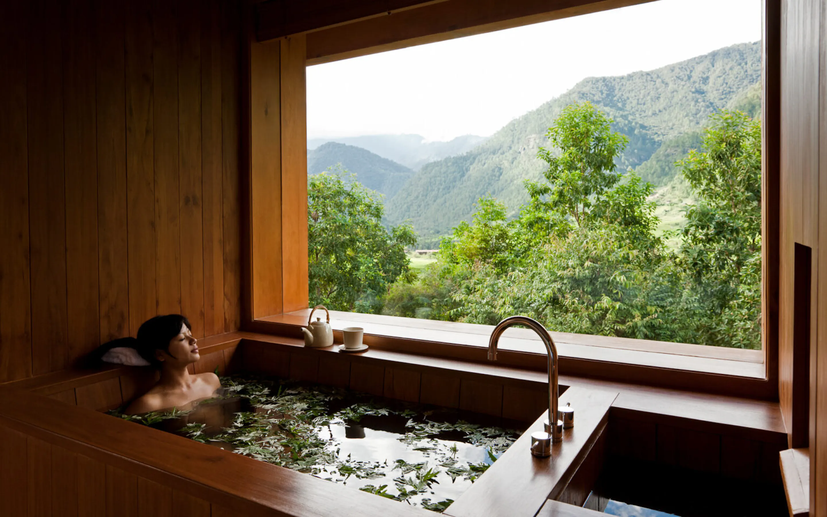 A woman relaxes in a deep tub at COMO Uma Punakha, framed by wood paneling and misty hills beyond the window.