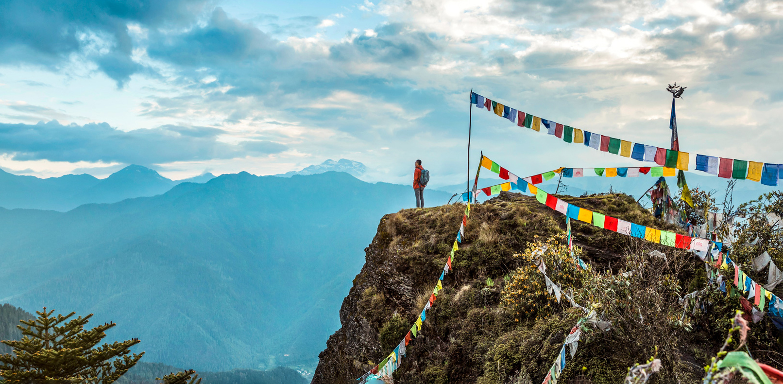 Prayer flags flutter from a rocky peak above COMO Uma Paro, with layers of blue mountains filling the distance.