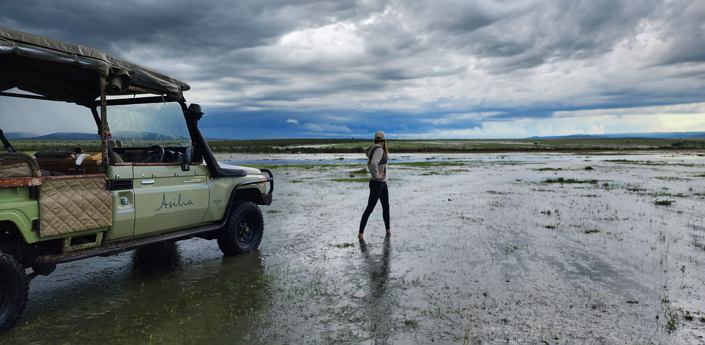 A traveler standing beside a safari vehicle on flooded plains against the backdrop of Kenya's Maasai Mara.