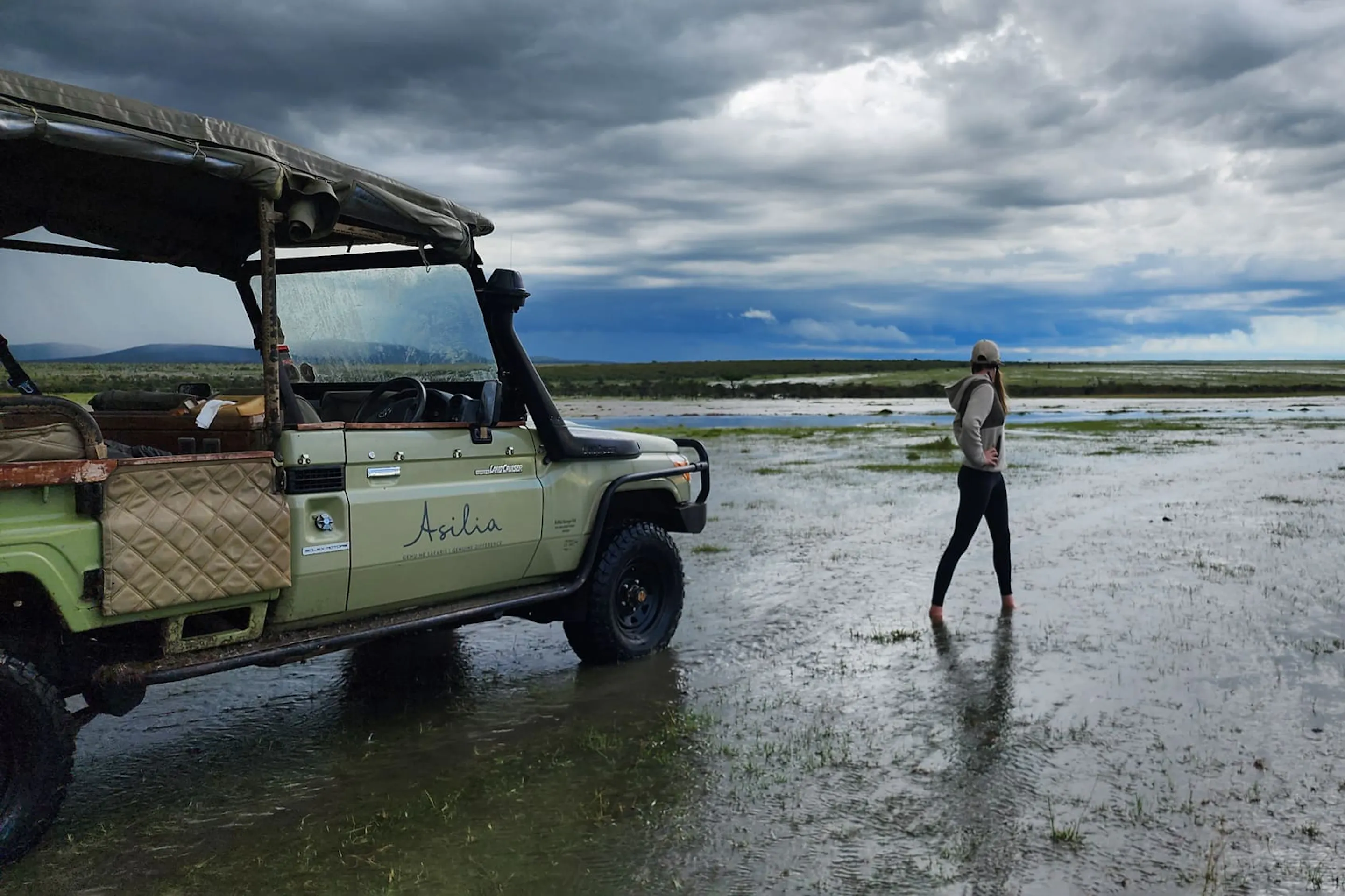 A traveler standing beside a safari vehicle on flooded plains against the backdrop of Kenya's Maasai Mara.