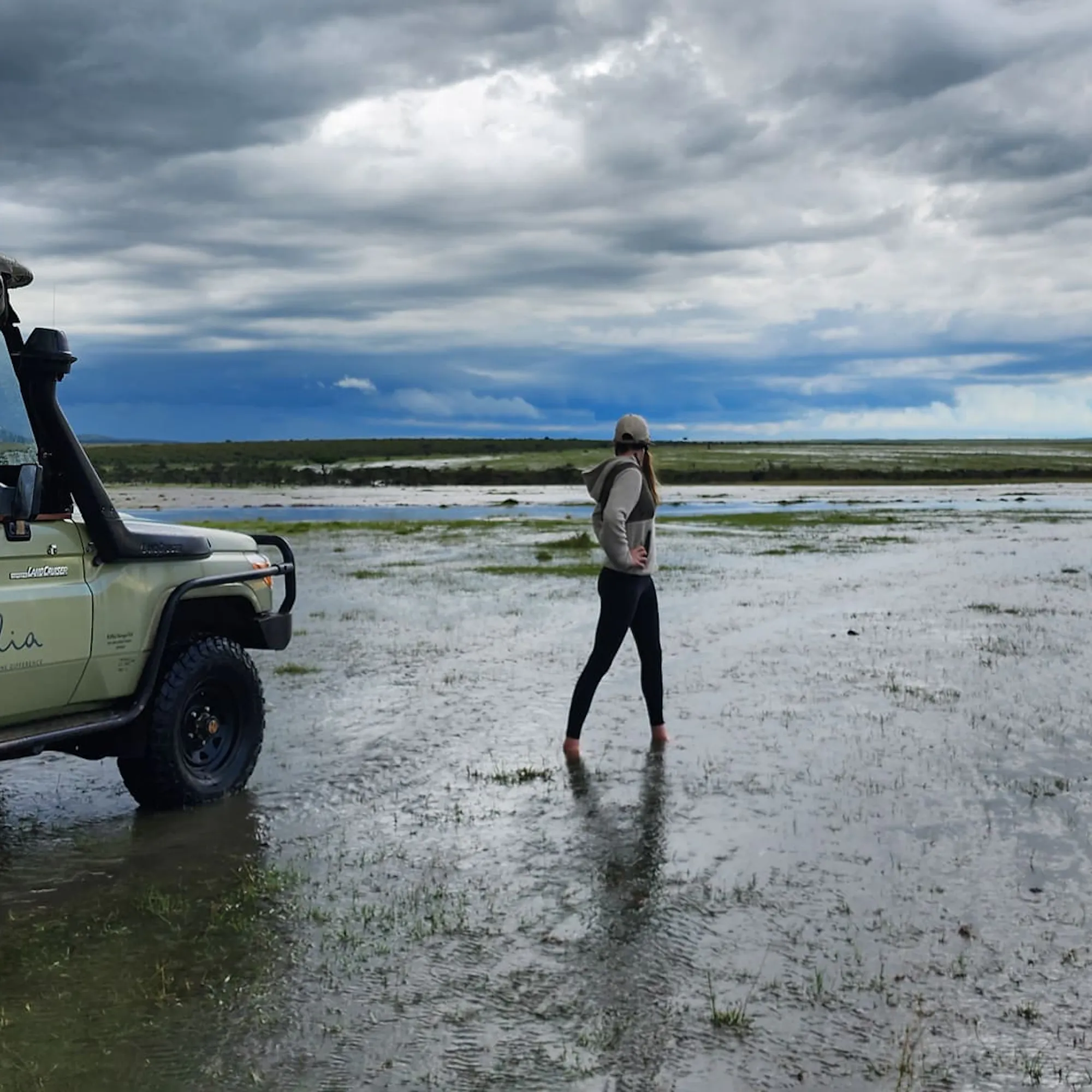 A traveler standing beside a safari vehicle on flooded plains against the backdrop of Kenya's Maasai Mara.