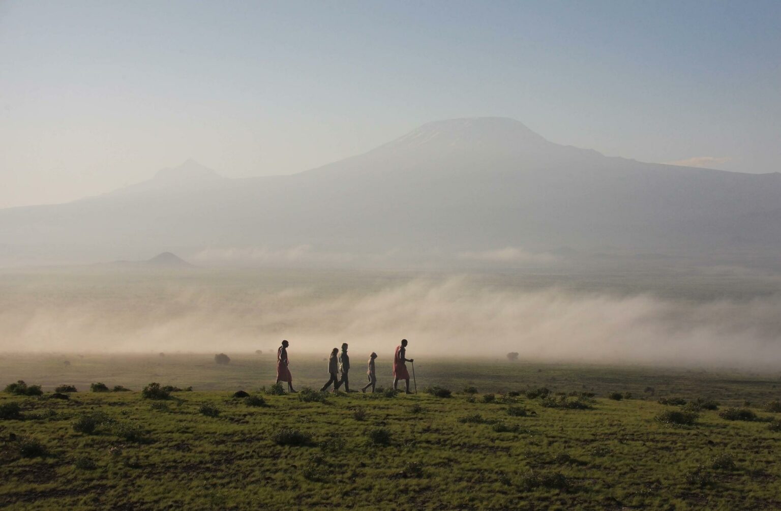 Five people walk across open grassland near Tortilis Camp in Amboseli, Kenya, with Mount Kilimanjaro in mist.