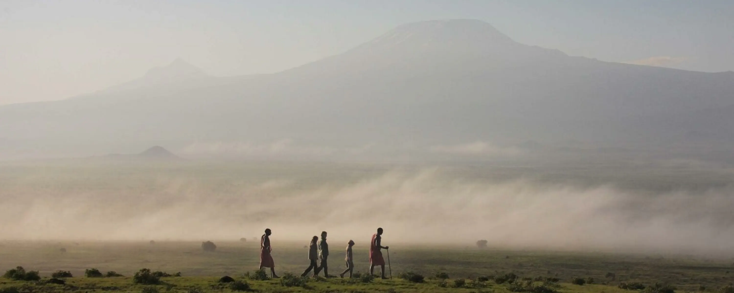 Five people walk across open grassland near Tortilis Camp in Amboseli, Kenya, with Mount Kilimanjaro in mist.