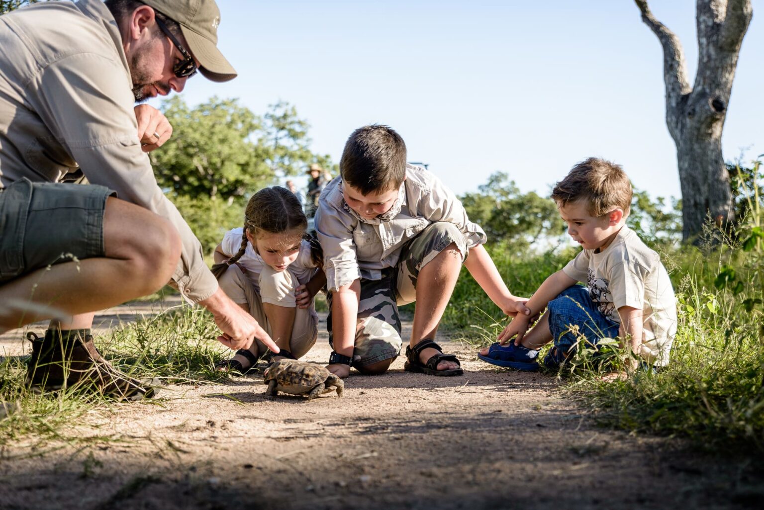 A guide crouches beside two children during a bush walk in Sabi Sand, showing them a bow and arrow in the grass.