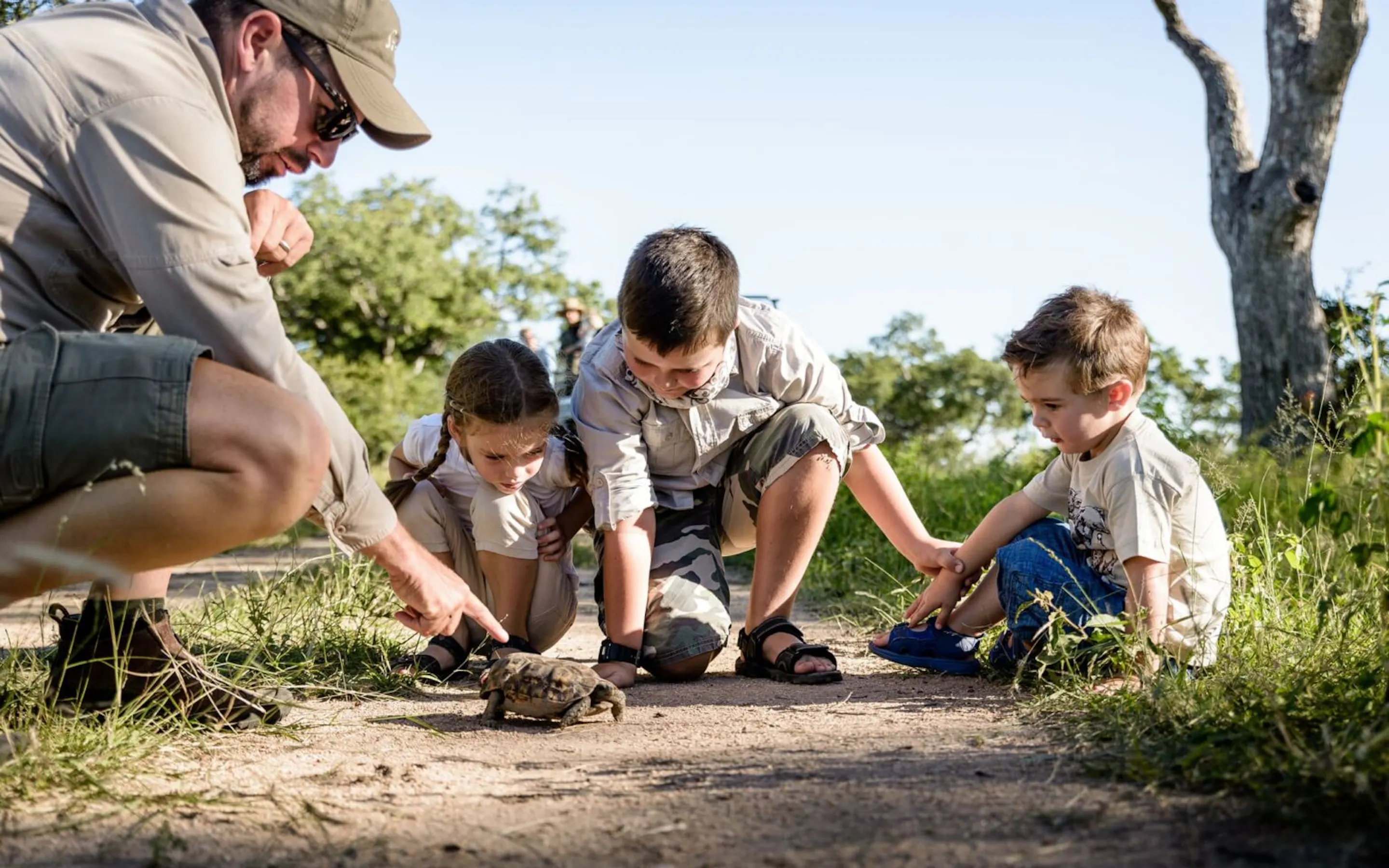 A guide crouches beside two children during a bush walk in Sabi Sand, showing them a bow and arrow in the grass.