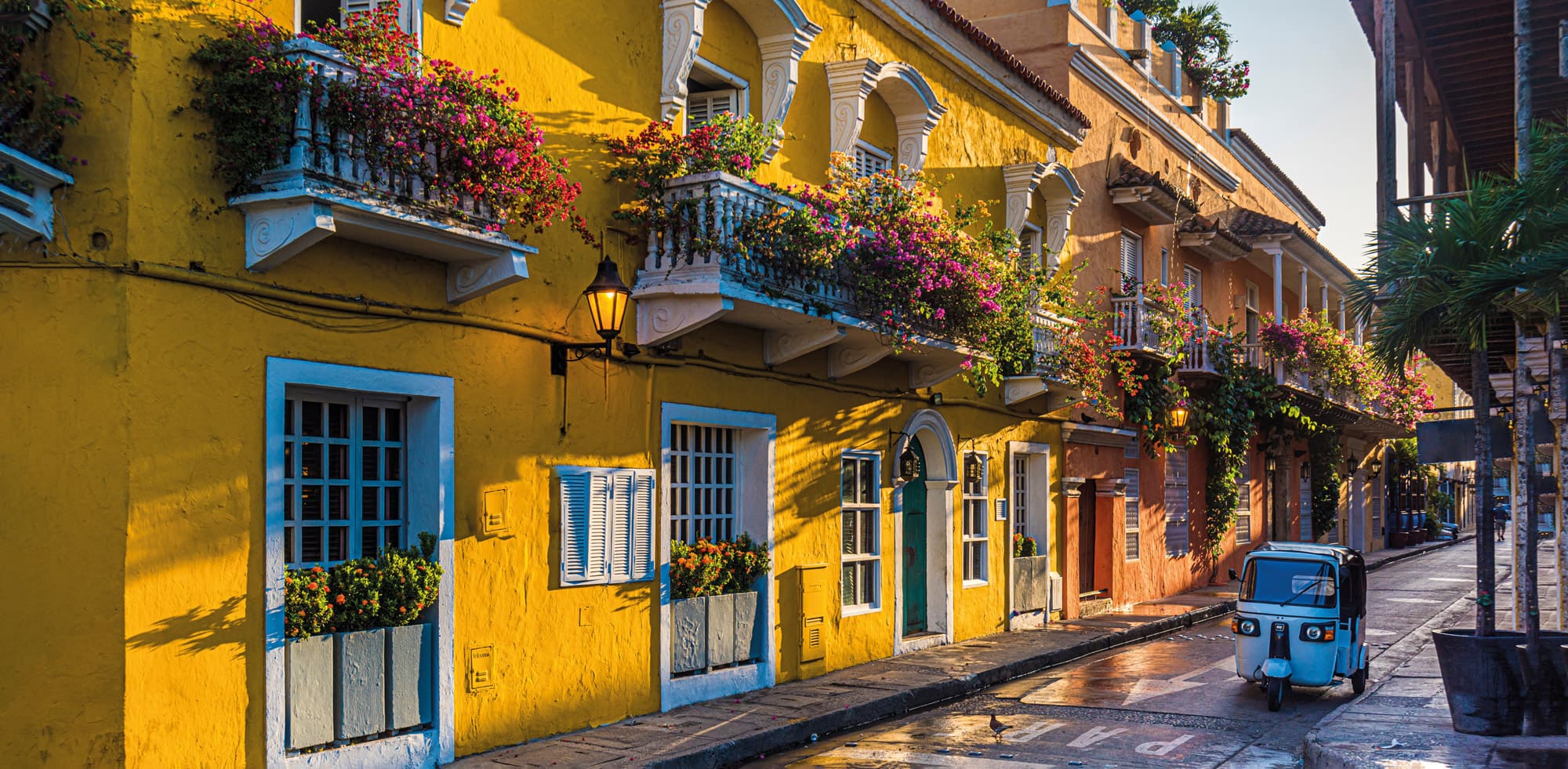 Colorful balconies and flower boxes line a narrow street in Cartagena's old town, lit by warm afternoon sun.