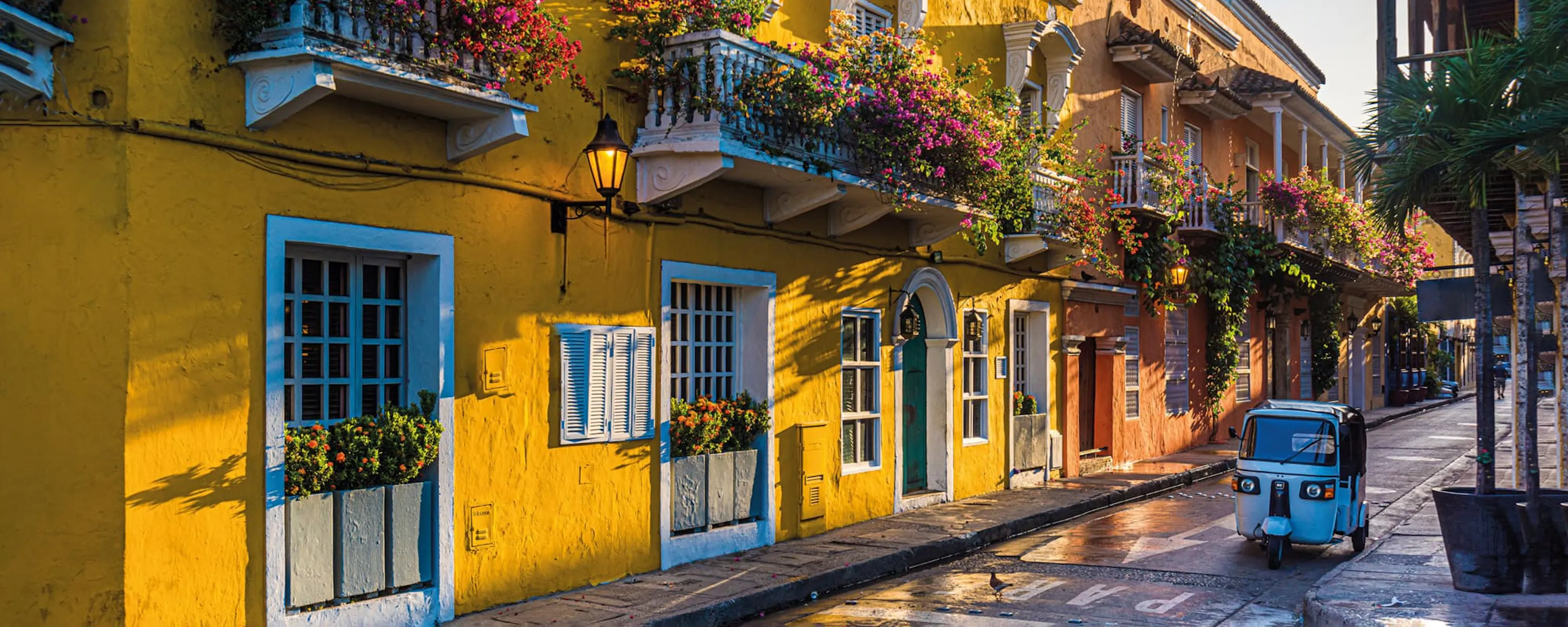 Colorful balconies and flower boxes line a narrow street in Cartagena's old town, lit by warm afternoon sun.
