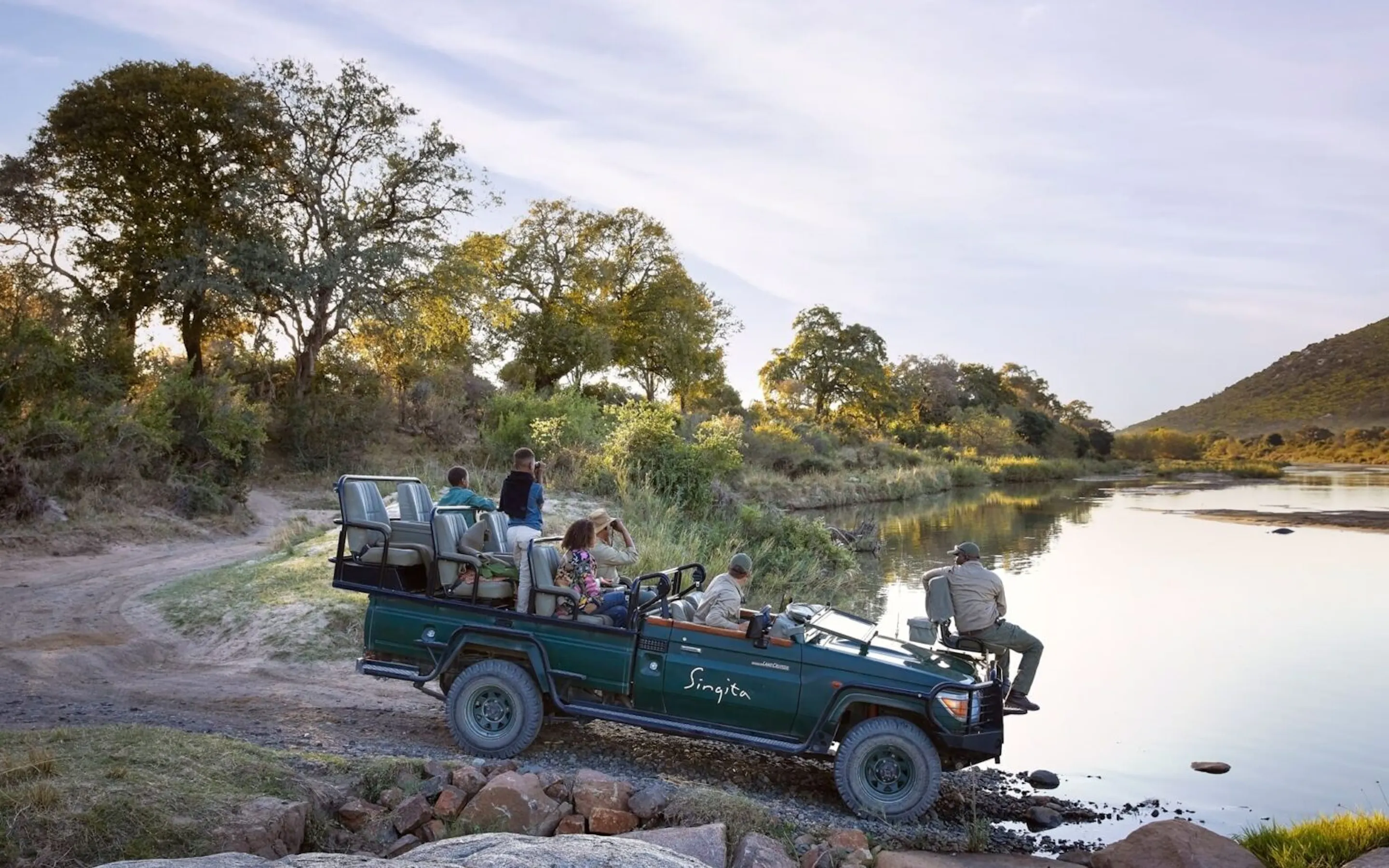 A family watches animals at the edges of a river, from an open safari vehicle in Sabi Sand