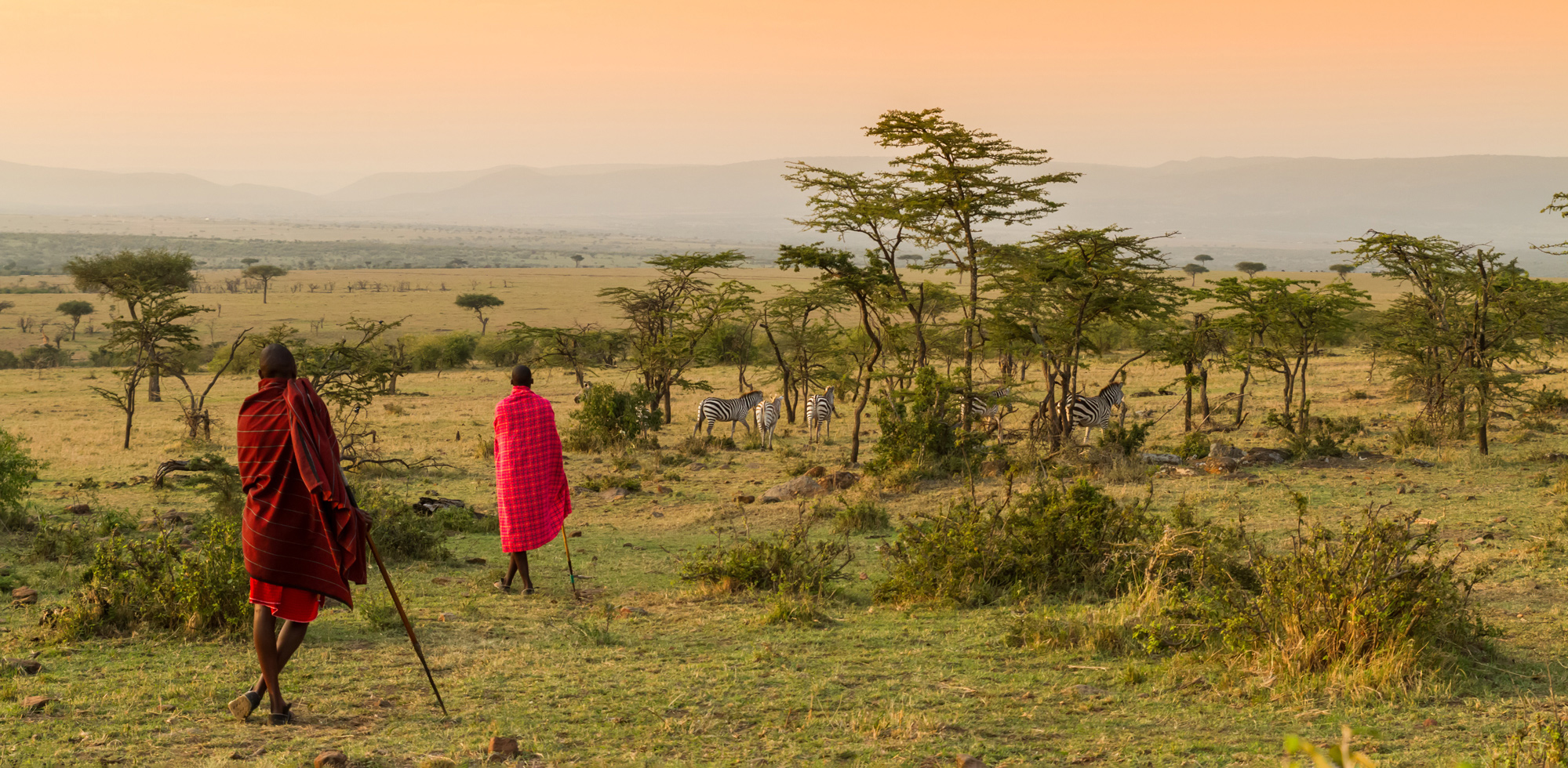 Two Maasai stand above Kenya's open plains, looking across golden grass under a dramatic sky near the Mara.