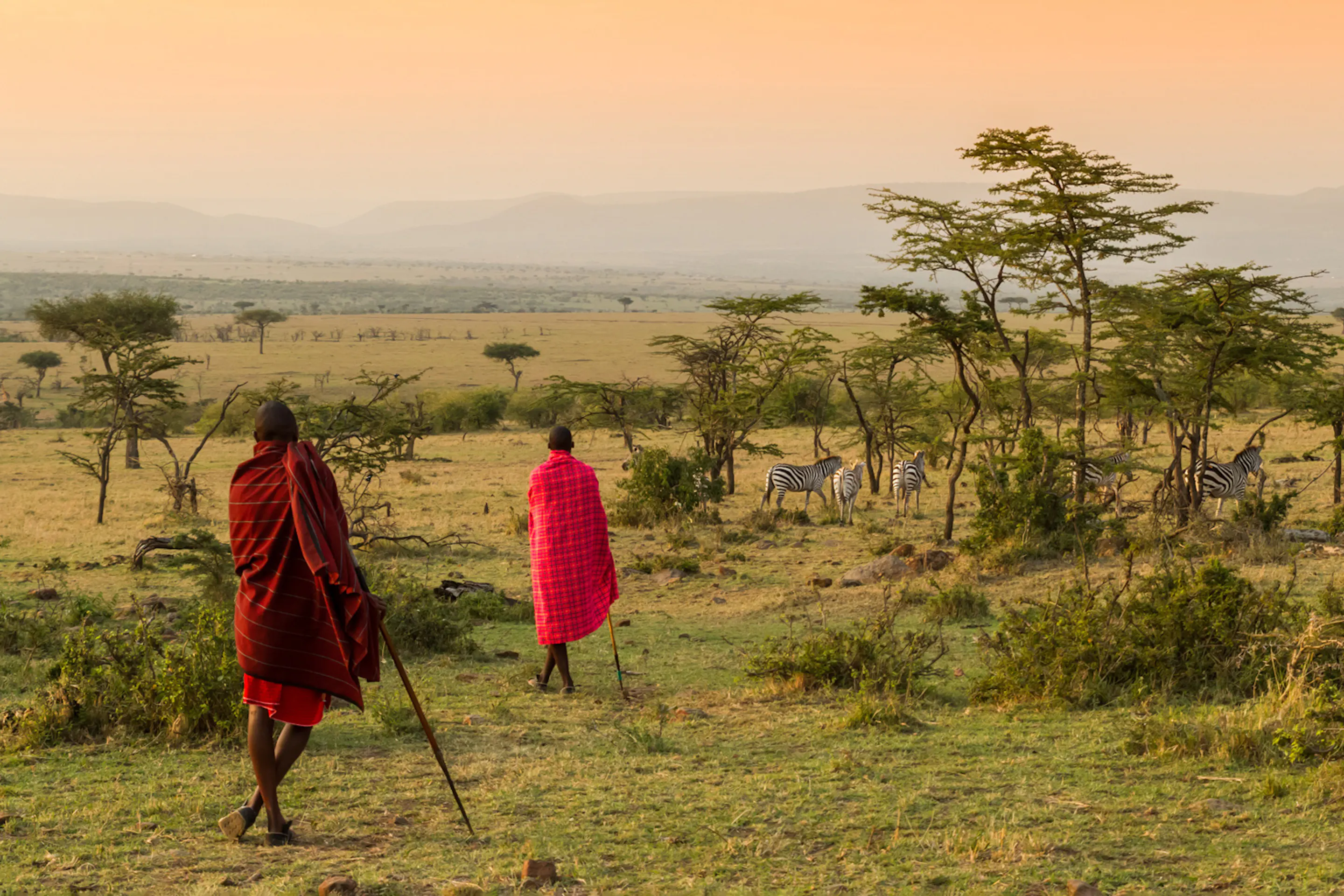 Two Maasai stand above Kenya's open plains, looking across golden grass under a dramatic sky near the Mara.