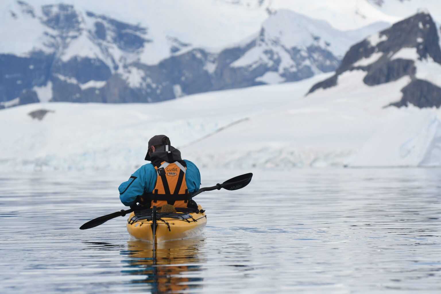 A kayaker crossing icy Antarctic waters beneath snowy peaks against the backdrop of the Antarctic Peninsula.